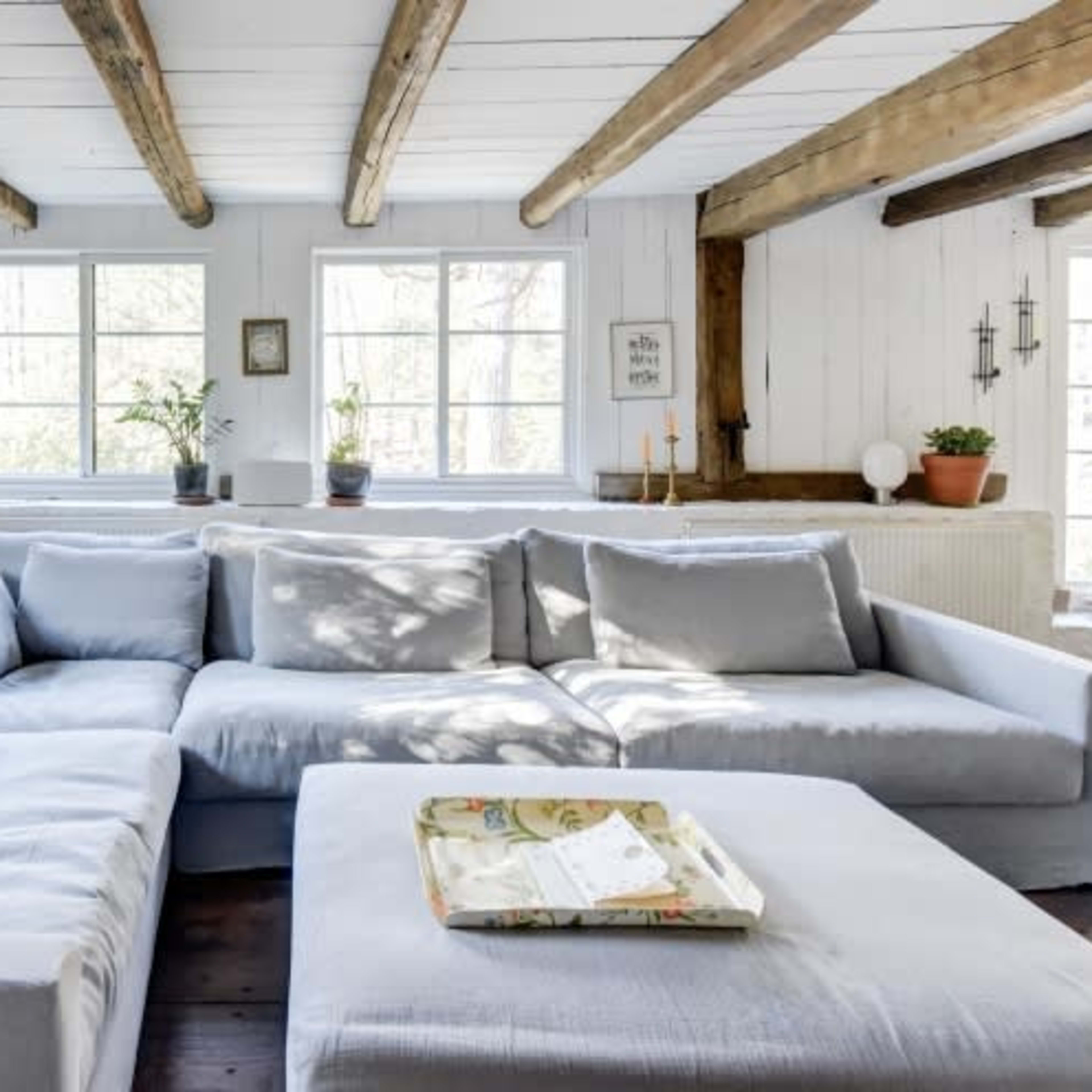 A bright, inviting living room featuring a large light gray sectional sofa, a coffee table, and several plants in windows.