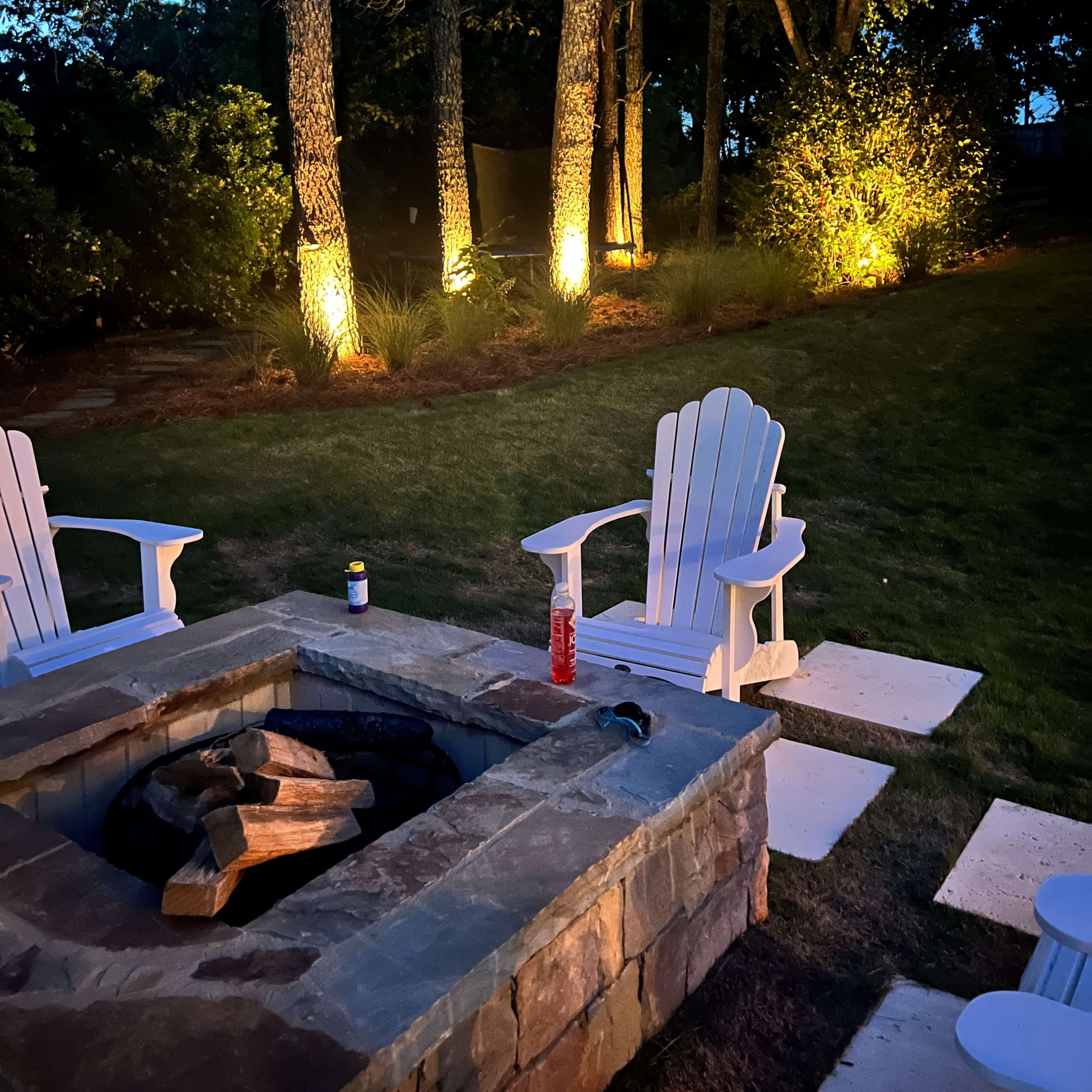 A stone fire pit surrounded by two white Adirondack chairs in a well-lit backyard at dusk.
