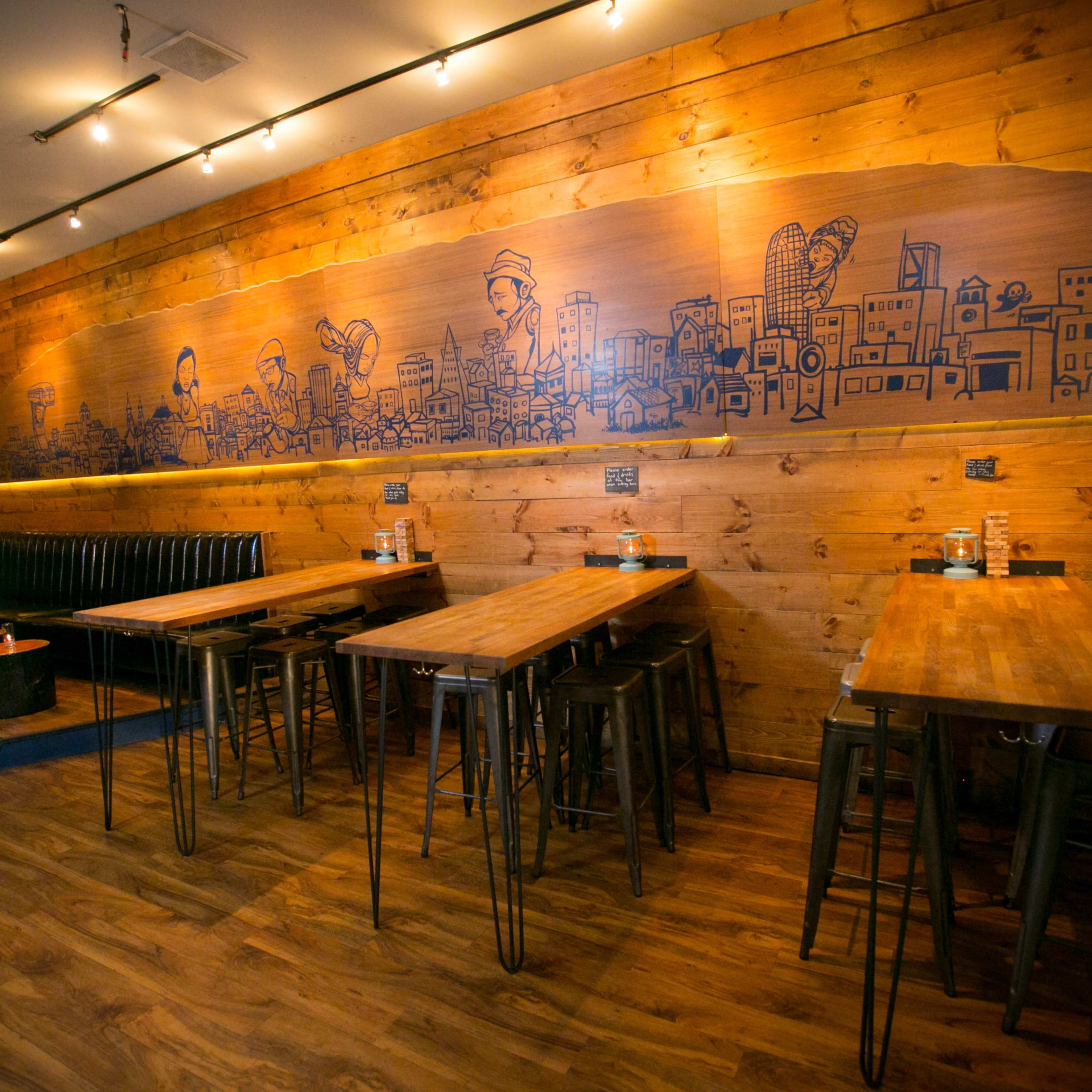 A wooden-themed restaurant interior with multiple wooden tables, tall stools, and a mural depicting a cityscape on the wall.