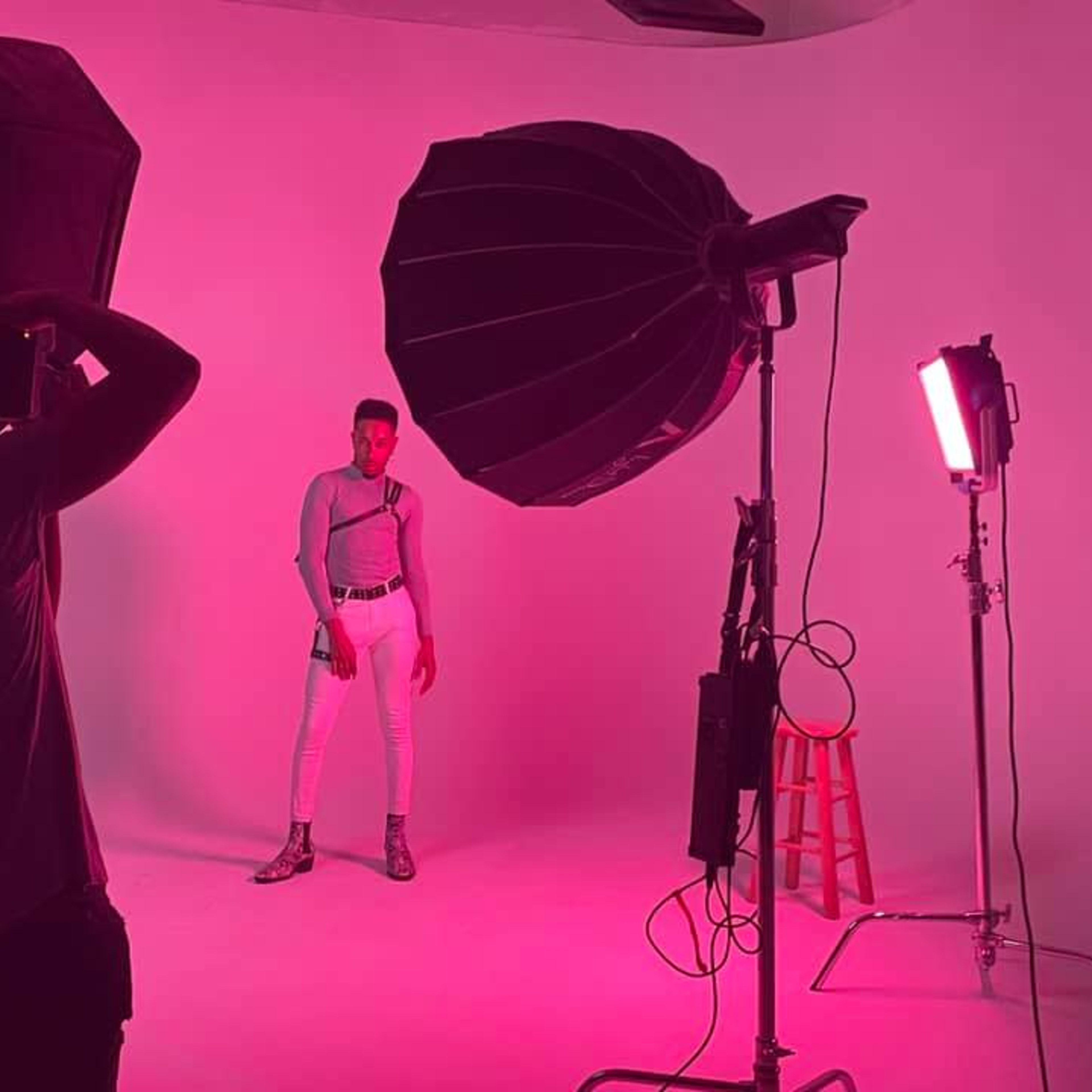 A model poses in front of a pink backdrop while a photographer captures the shot under studio lights.