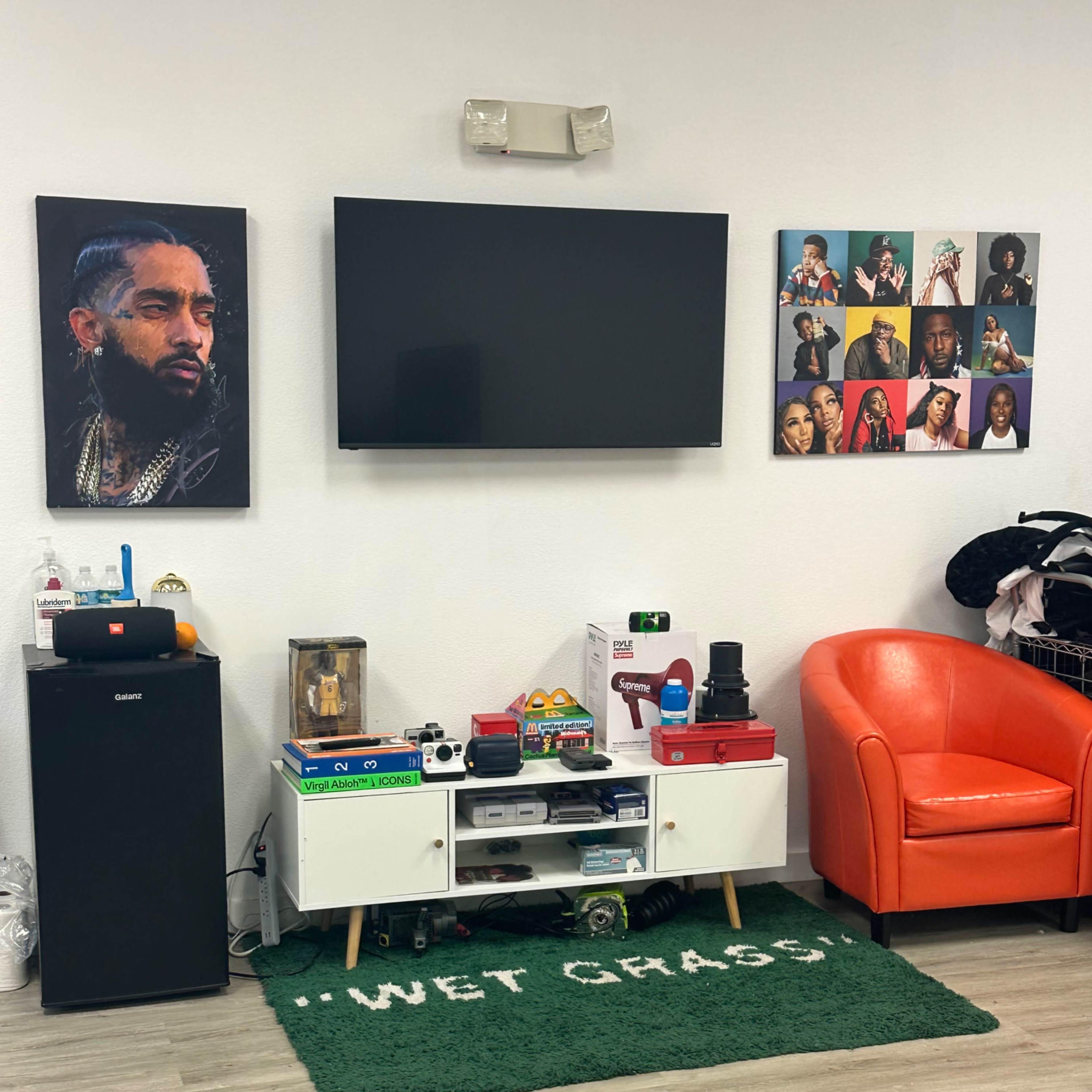 A room with a large flat-screen TV mounted on the wall, a black mini-fridge, a white entertainment unit filled with various items, an orange chair, and several framed pictures of musicians displayed on the wall.