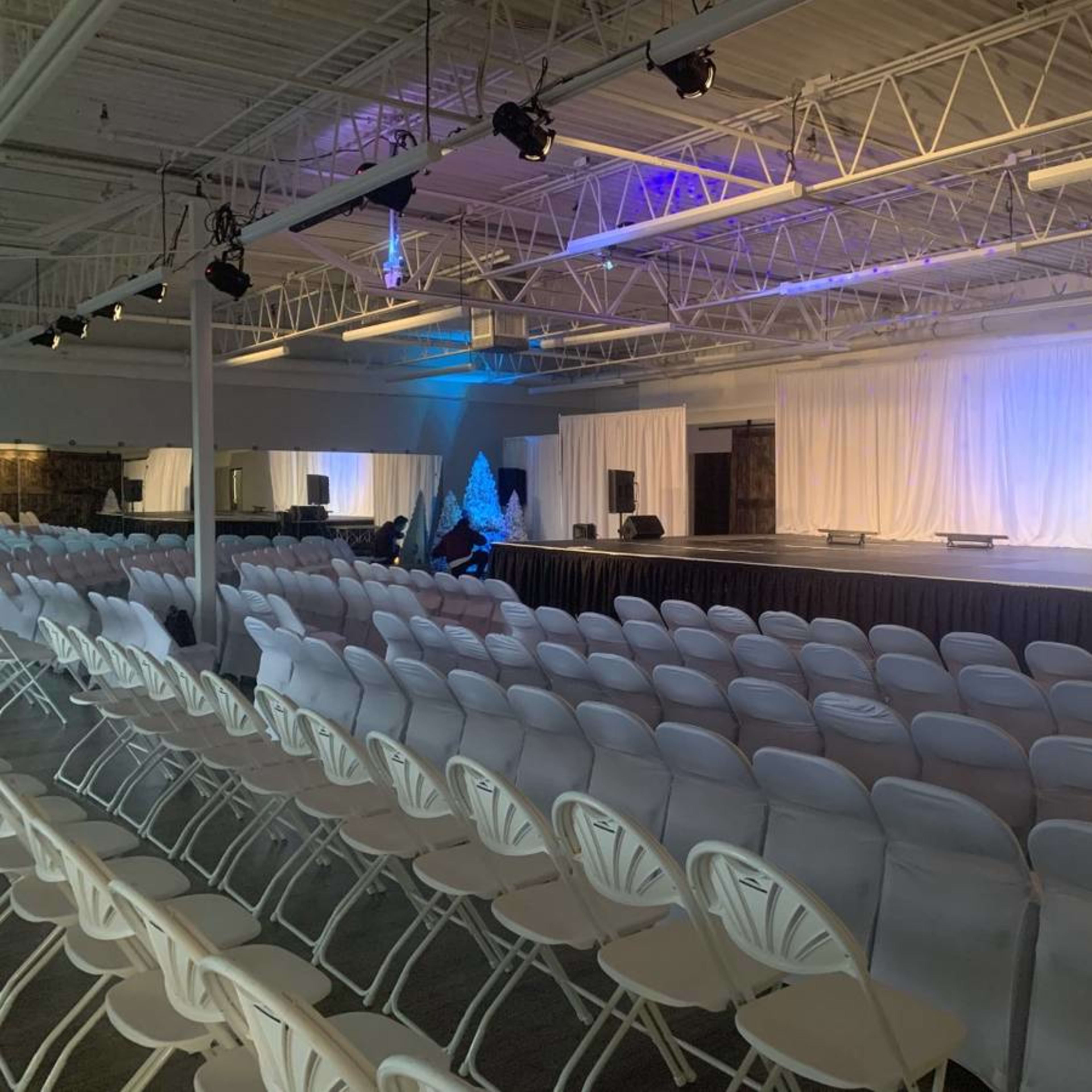 A spacious event hall set up with rows of white chairs facing a stage adorned with a white backdrop and dim lighting.