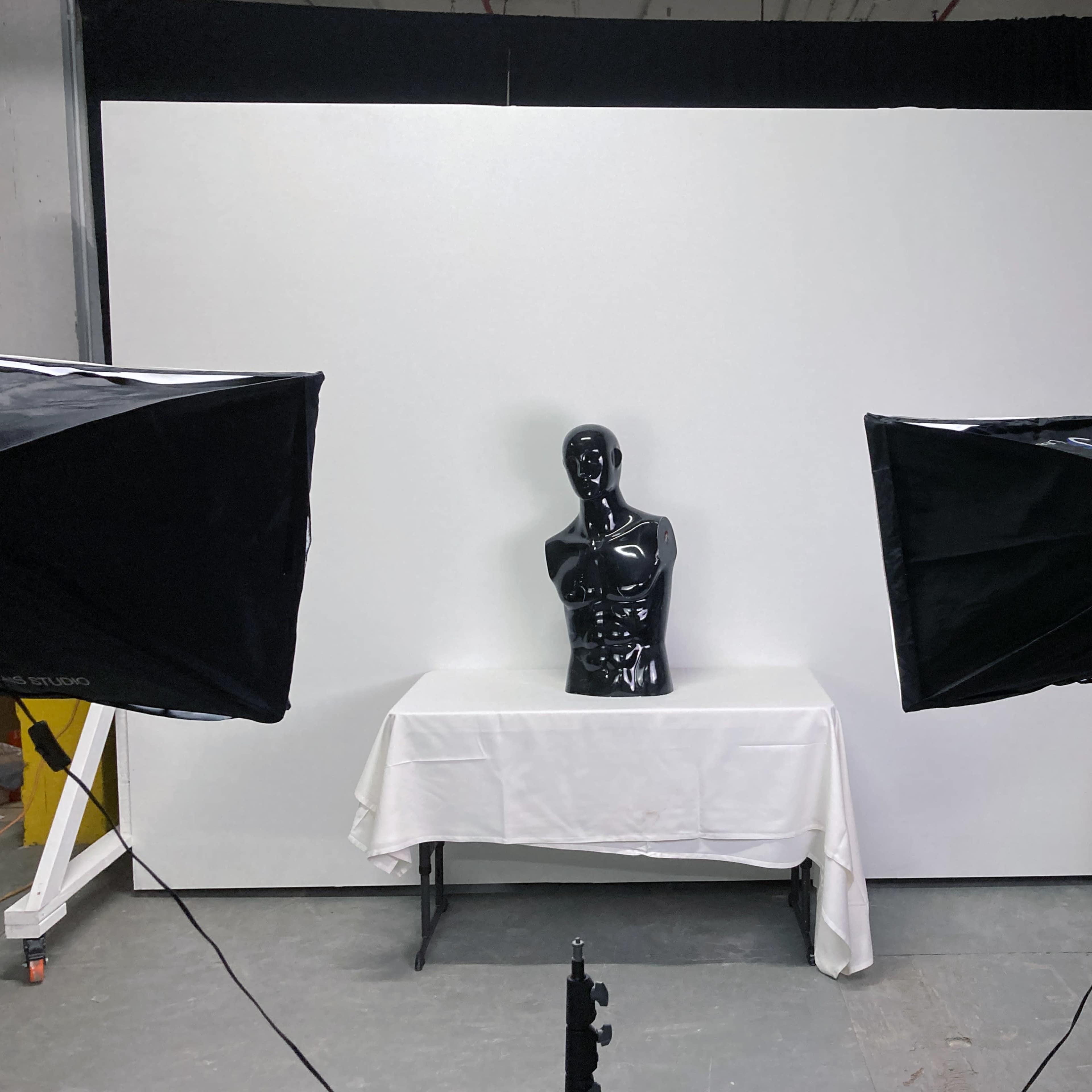 A black mannequin placed on a table covered with a white cloth, flanked by two large softbox lights, set against a plain white backdrop.