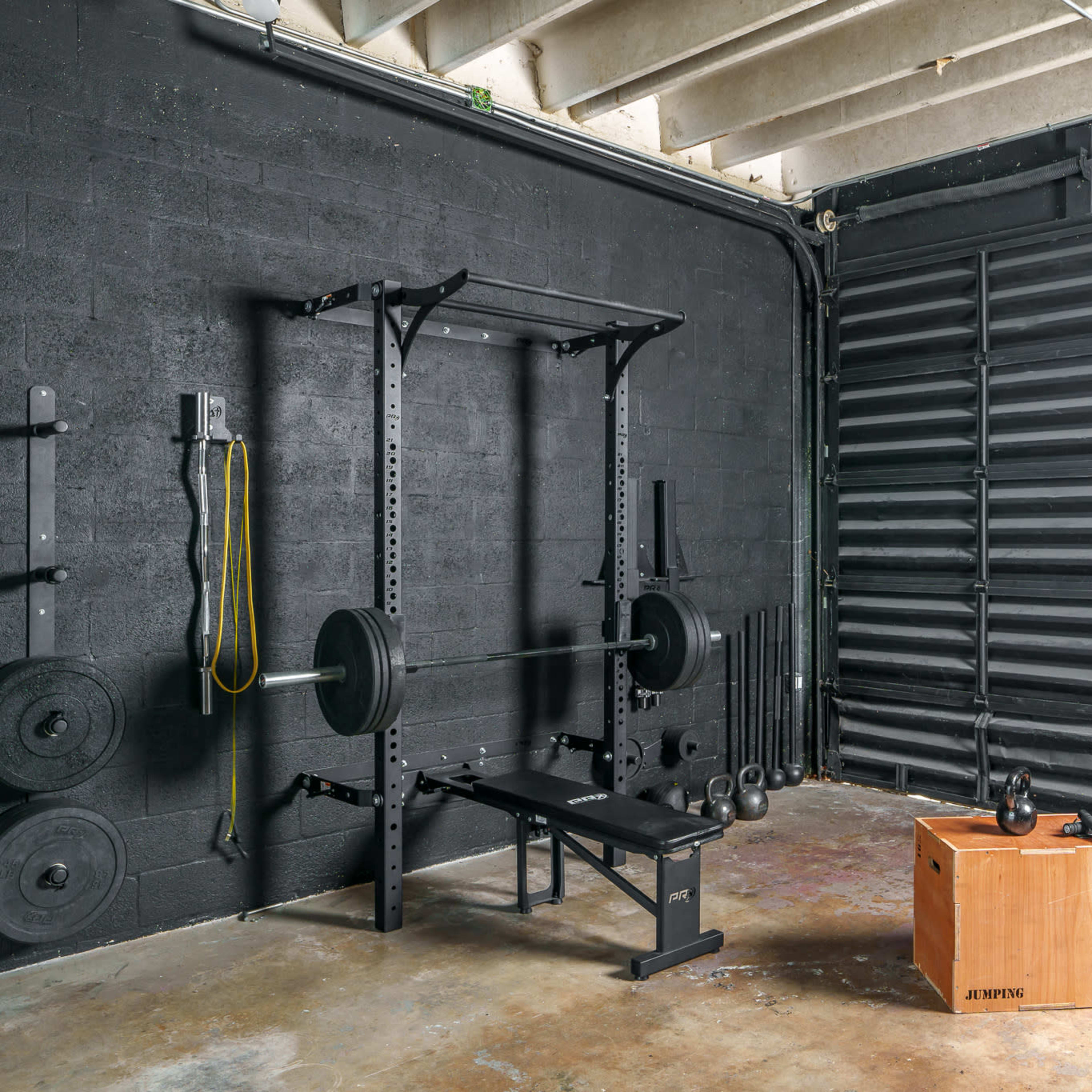 A gym setup in a black-walled room featuring a weightlifting rack with a barbell, a bench, a wooden plyometric box, a kettlebell, and a punching bag.