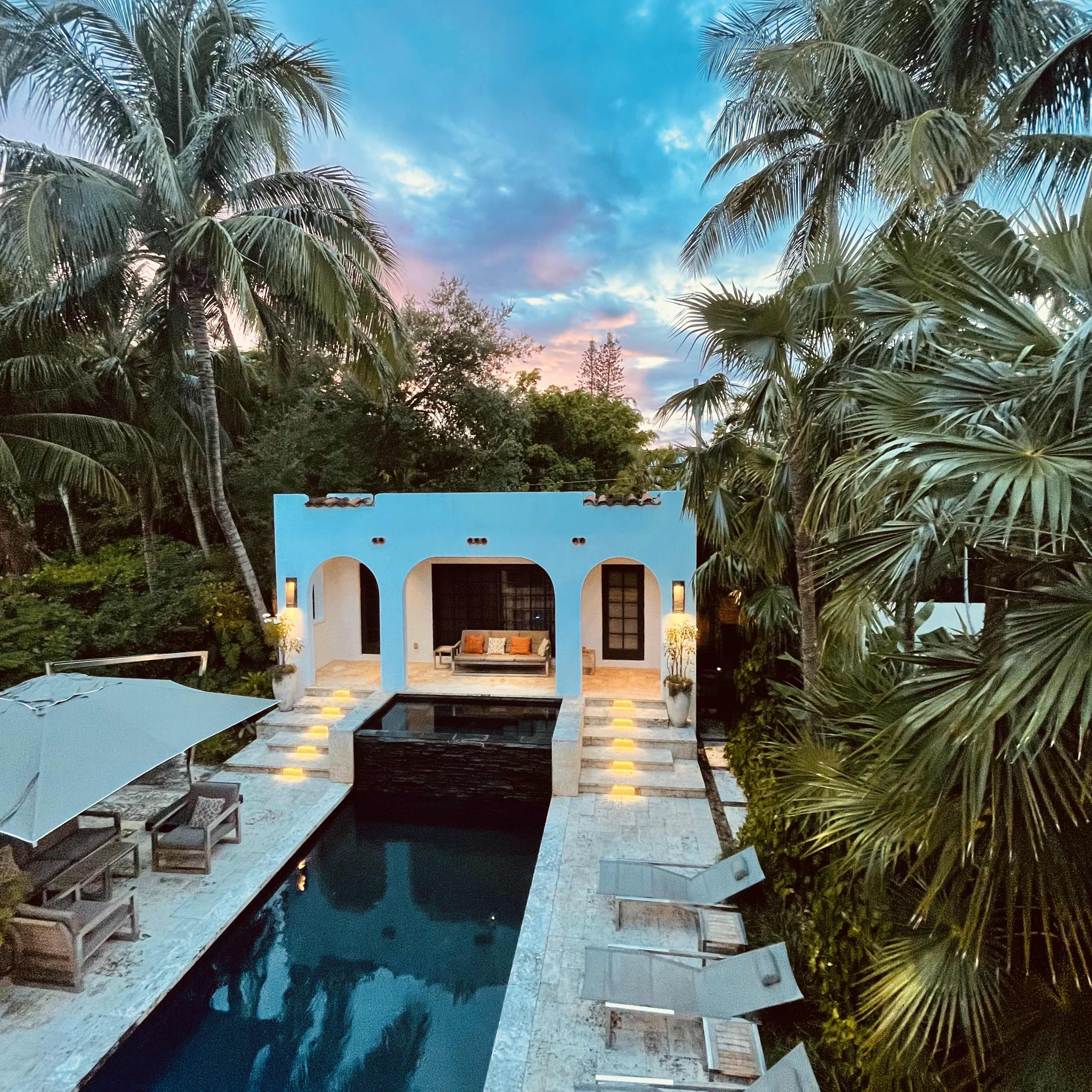 A pool surrounded by palm trees, with a blue building and lounge chairs nearby, illuminated by pathway lights at dusk.