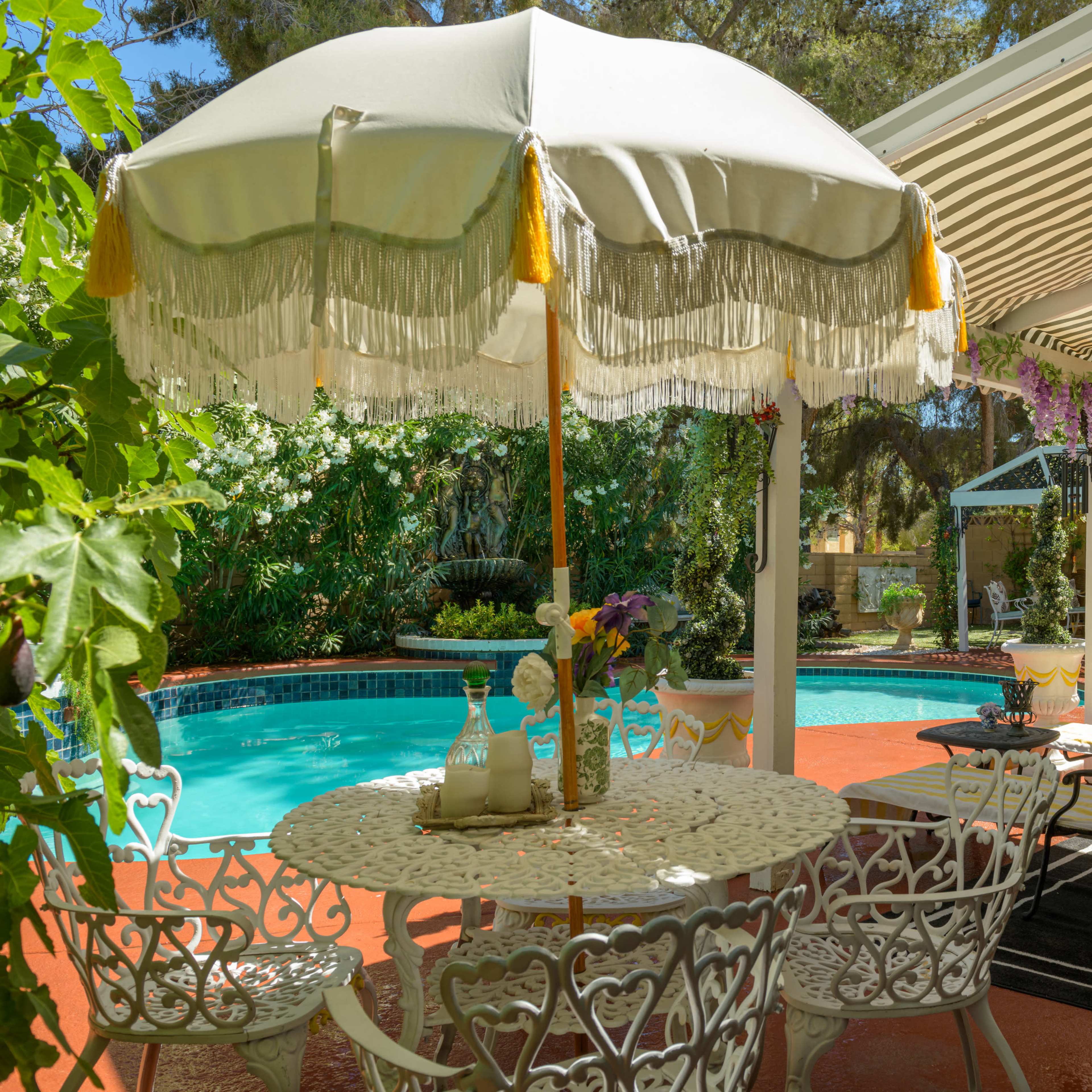 A shaded patio with a white umbrella overlooks a pool surrounded by lush greenery and floral accents.