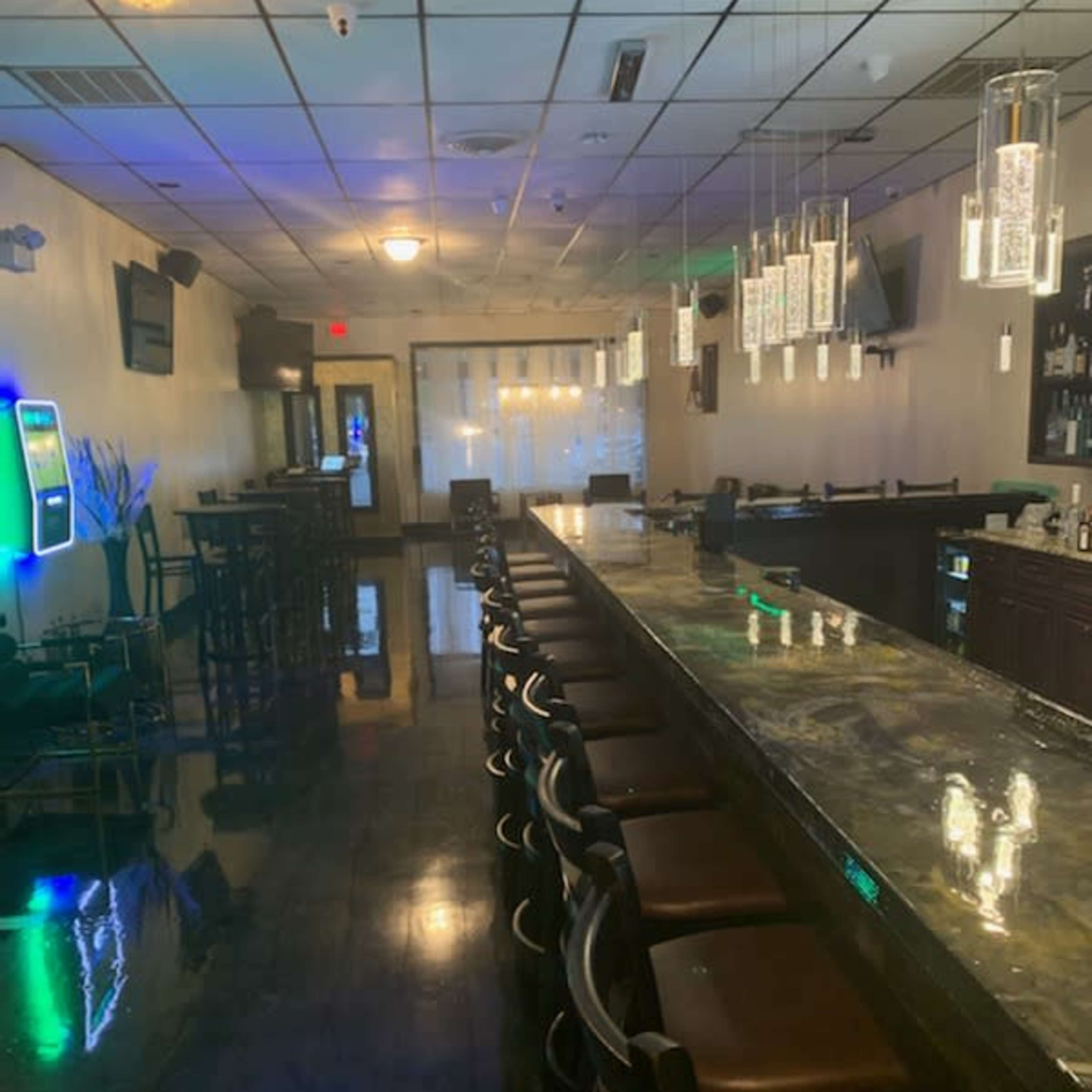 A long bar counter with chairs lined up in a dimly lit bar area, featuring a few hanging light fixtures and a distant view of a bar shelf.