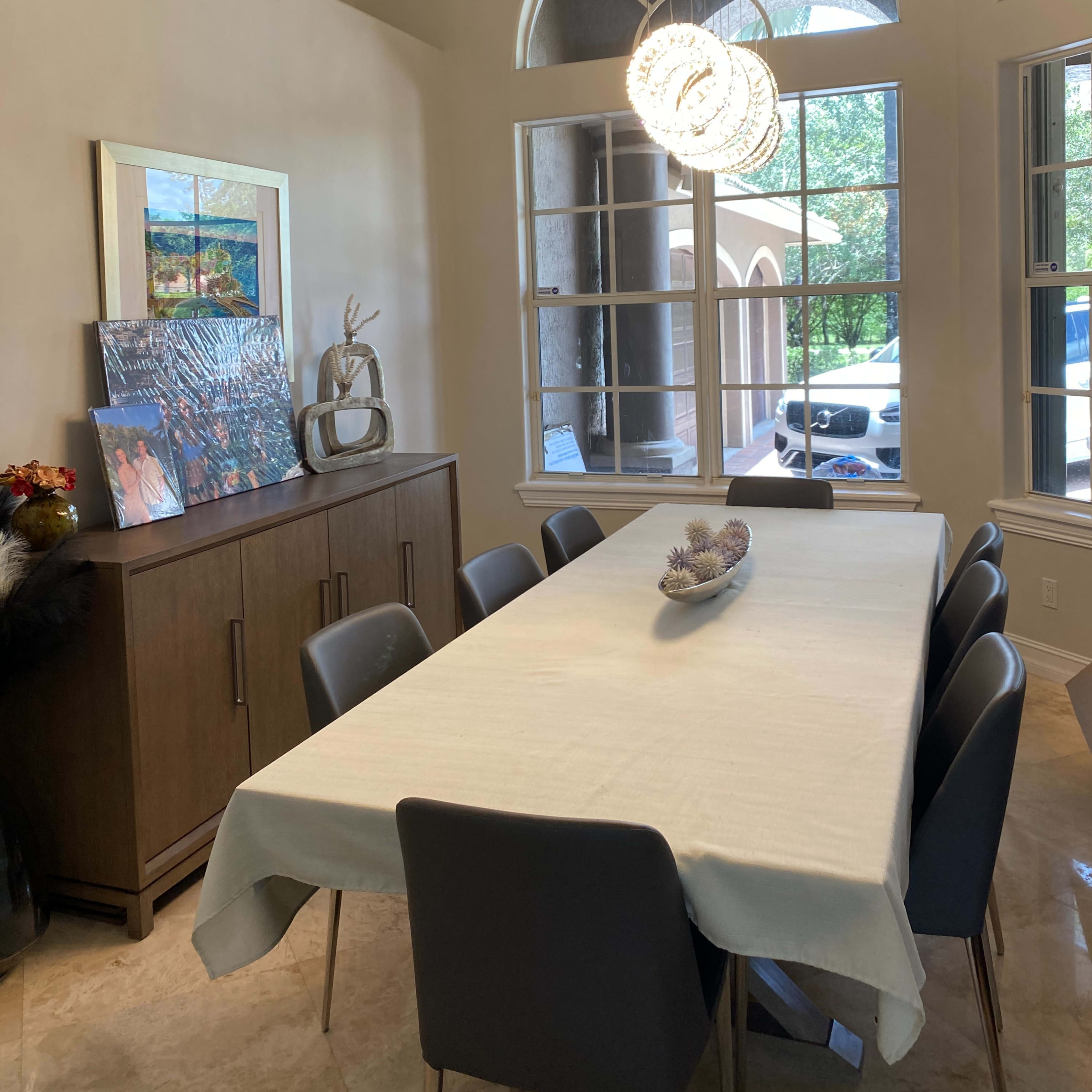 A long dining table is set in a well-lit room, flanked by modern chairs and large windows that allow natural light to enter.