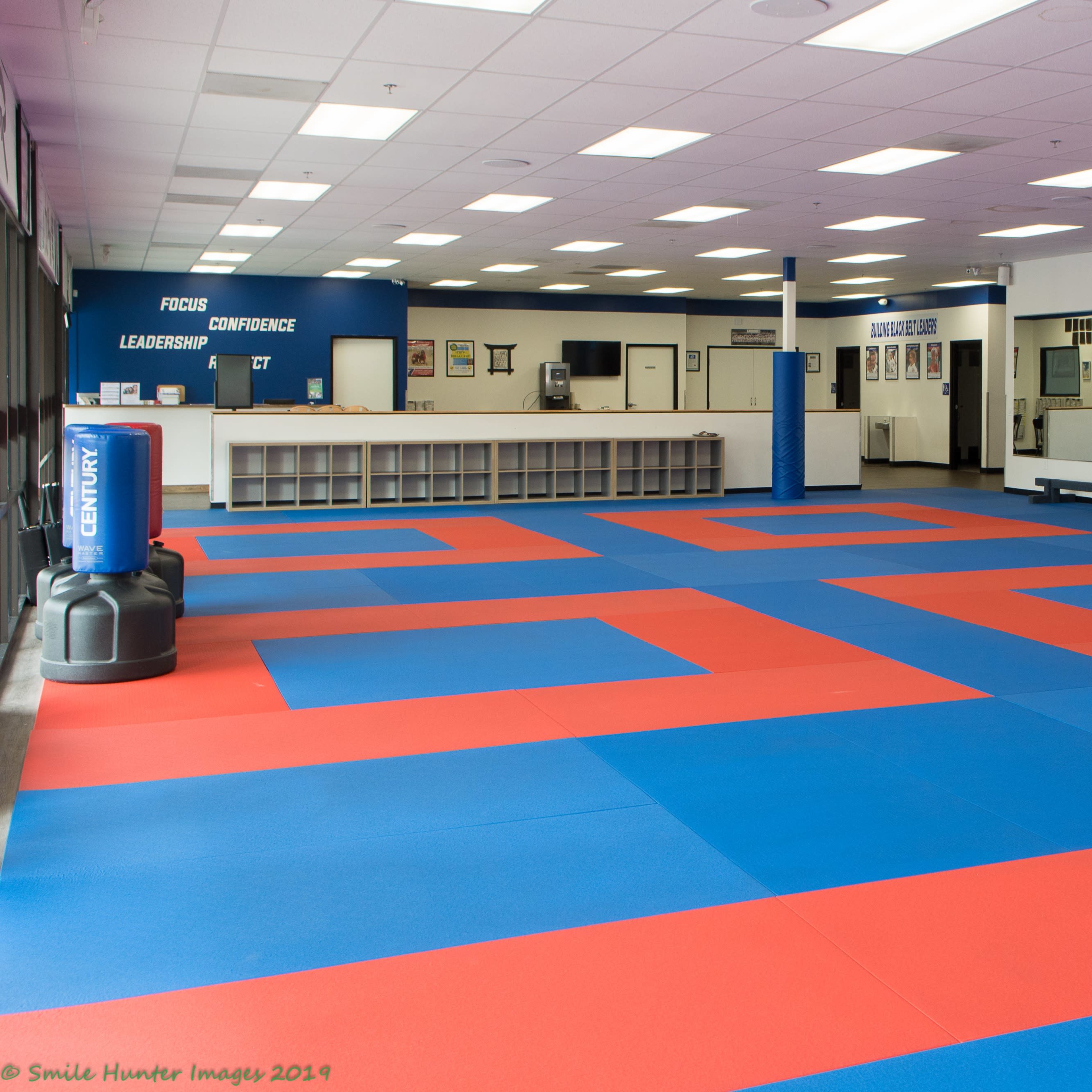 A spacious martial arts studio featuring red and blue mats, with a reception area and motivational signs on the walls.