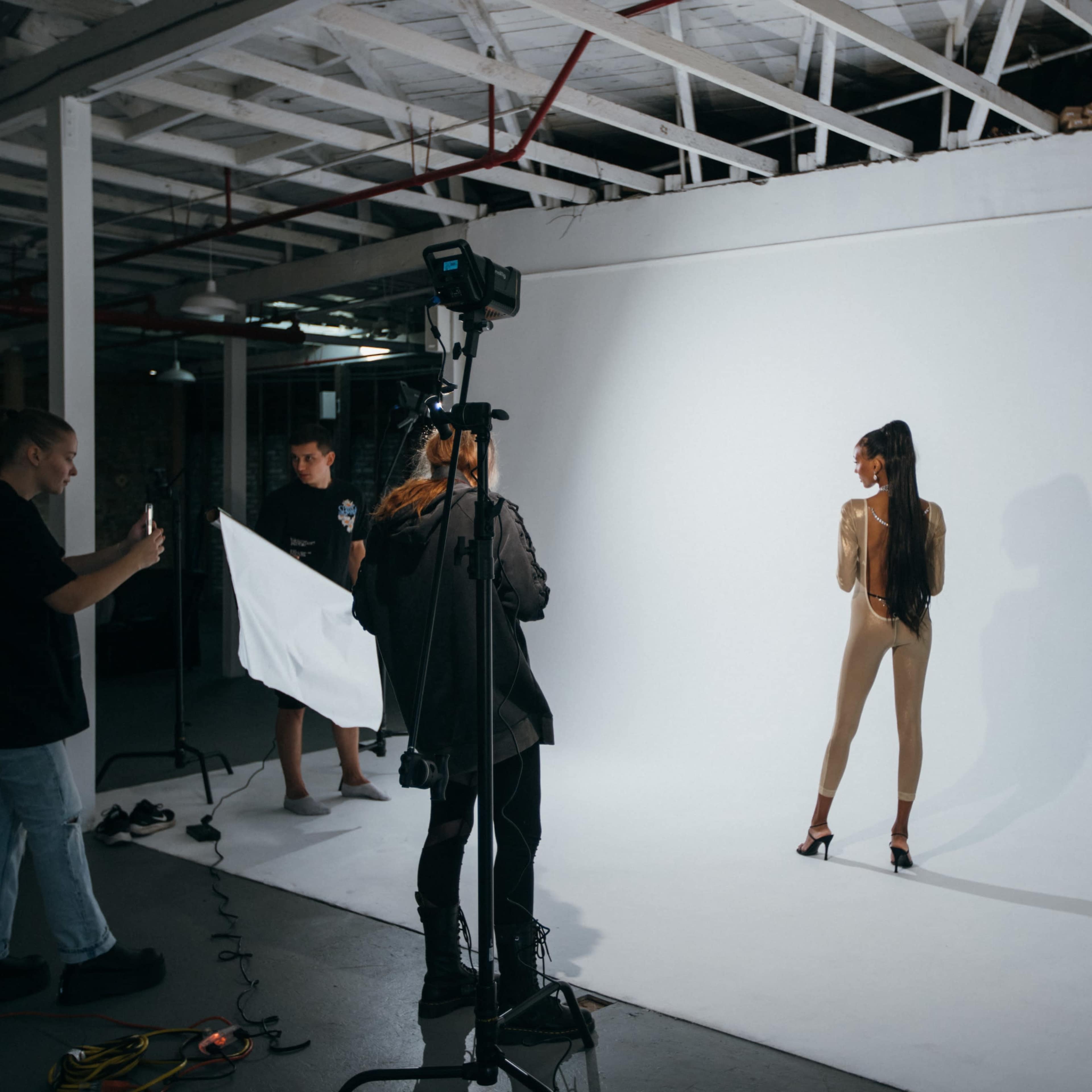 A model poses in front of a plain white backdrop while a small team sets up lighting and equipment in a studio.