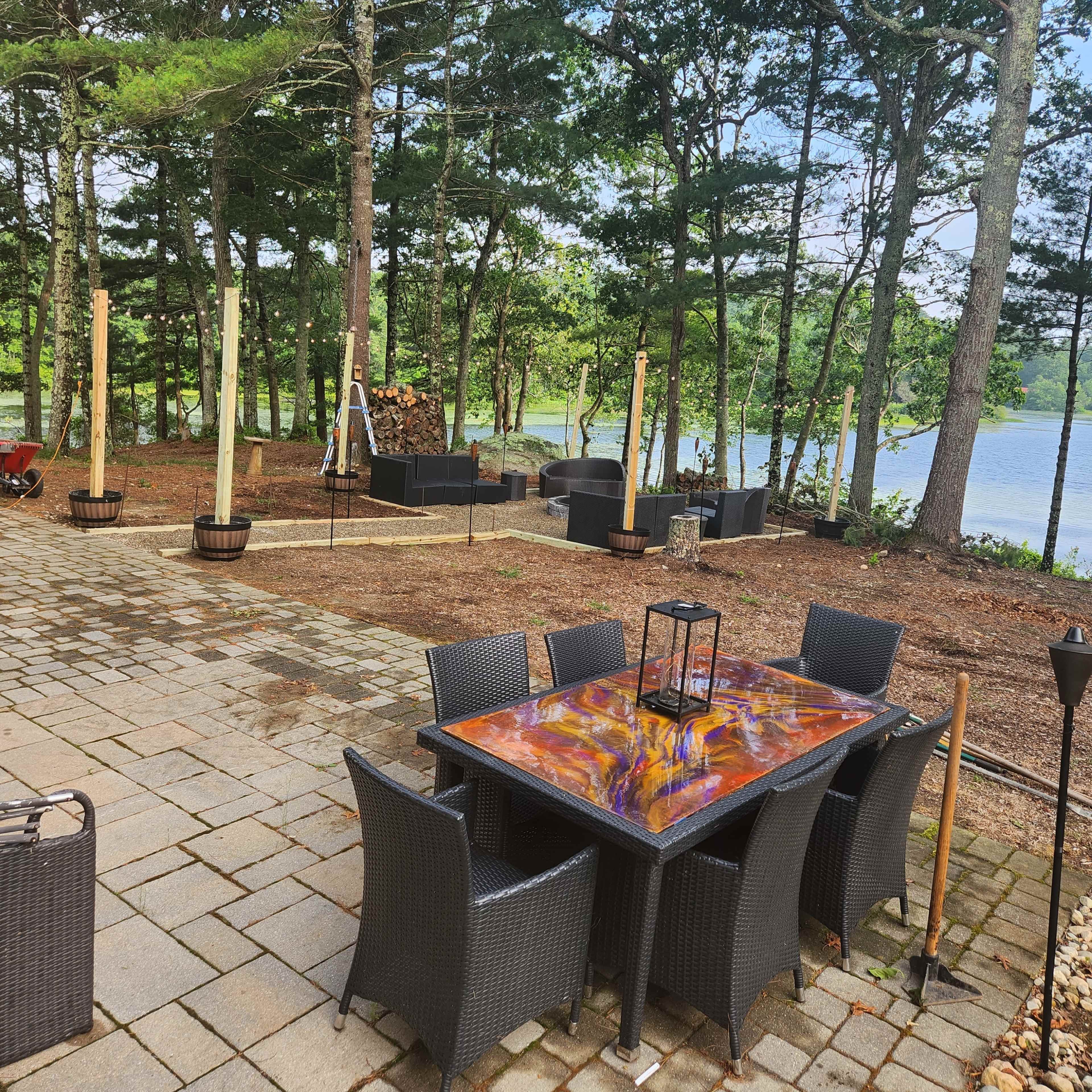 A patio area with a wooden table and chairs is set among trees near a lake, featuring stone pathways and various planters.
