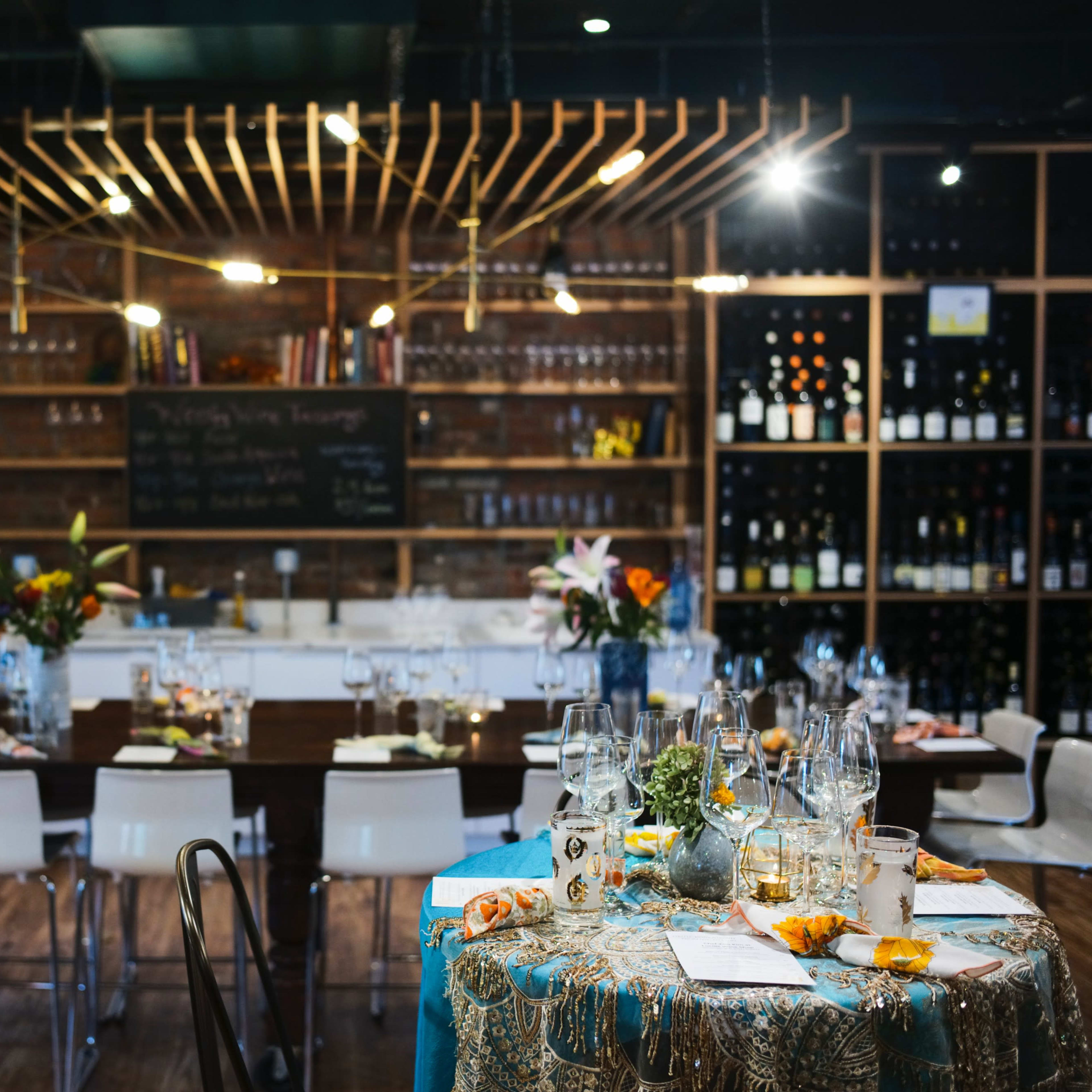 A dining area features a long wooden table set with glassware, surrounded by white chairs, and is decorated with flowers and an assortment of dishes, while a bar area displays shelves of wine.