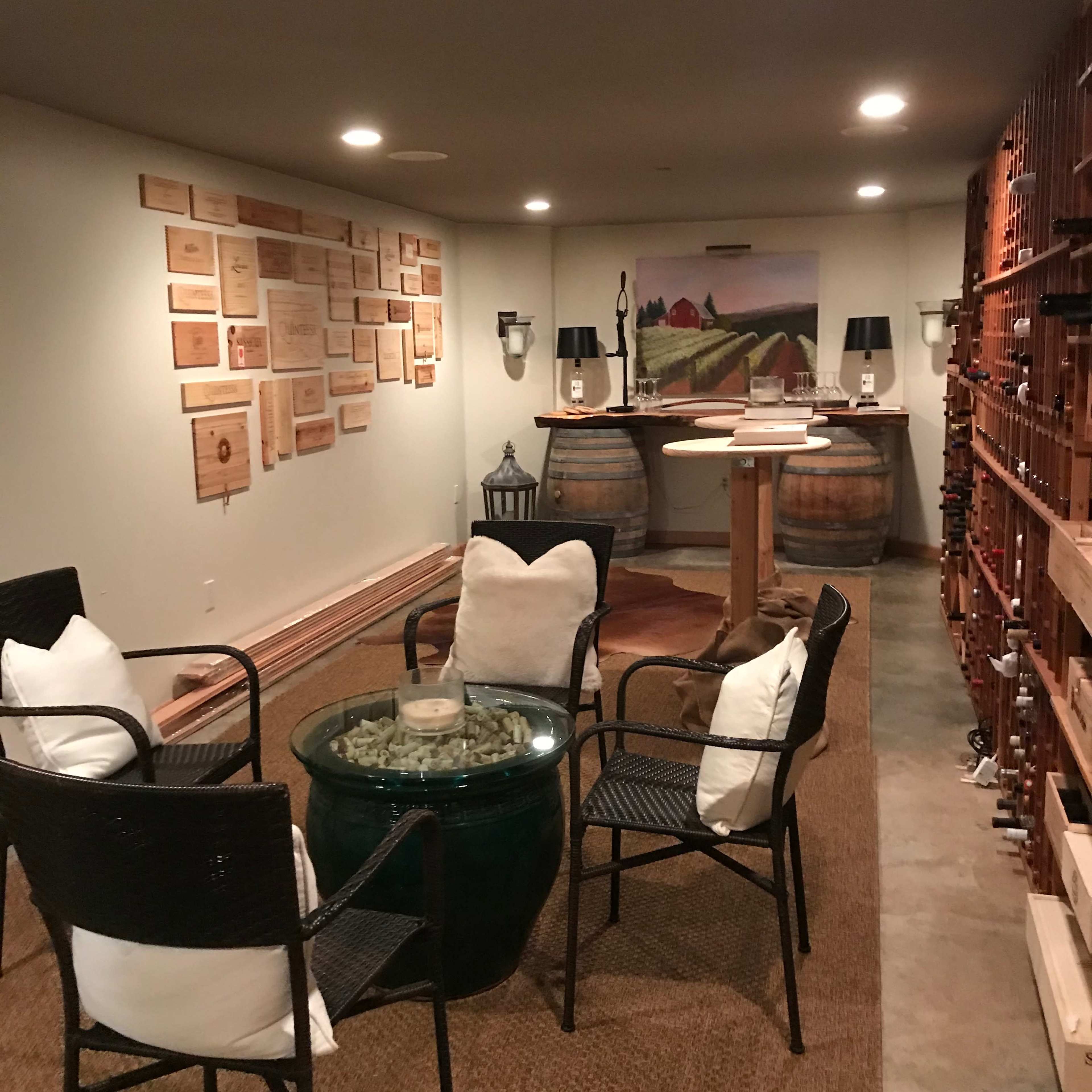 A cozy wine cellar featuring a seating area with four chairs surrounding a glass table, wooden barrels, and wine racks lined along the walls.