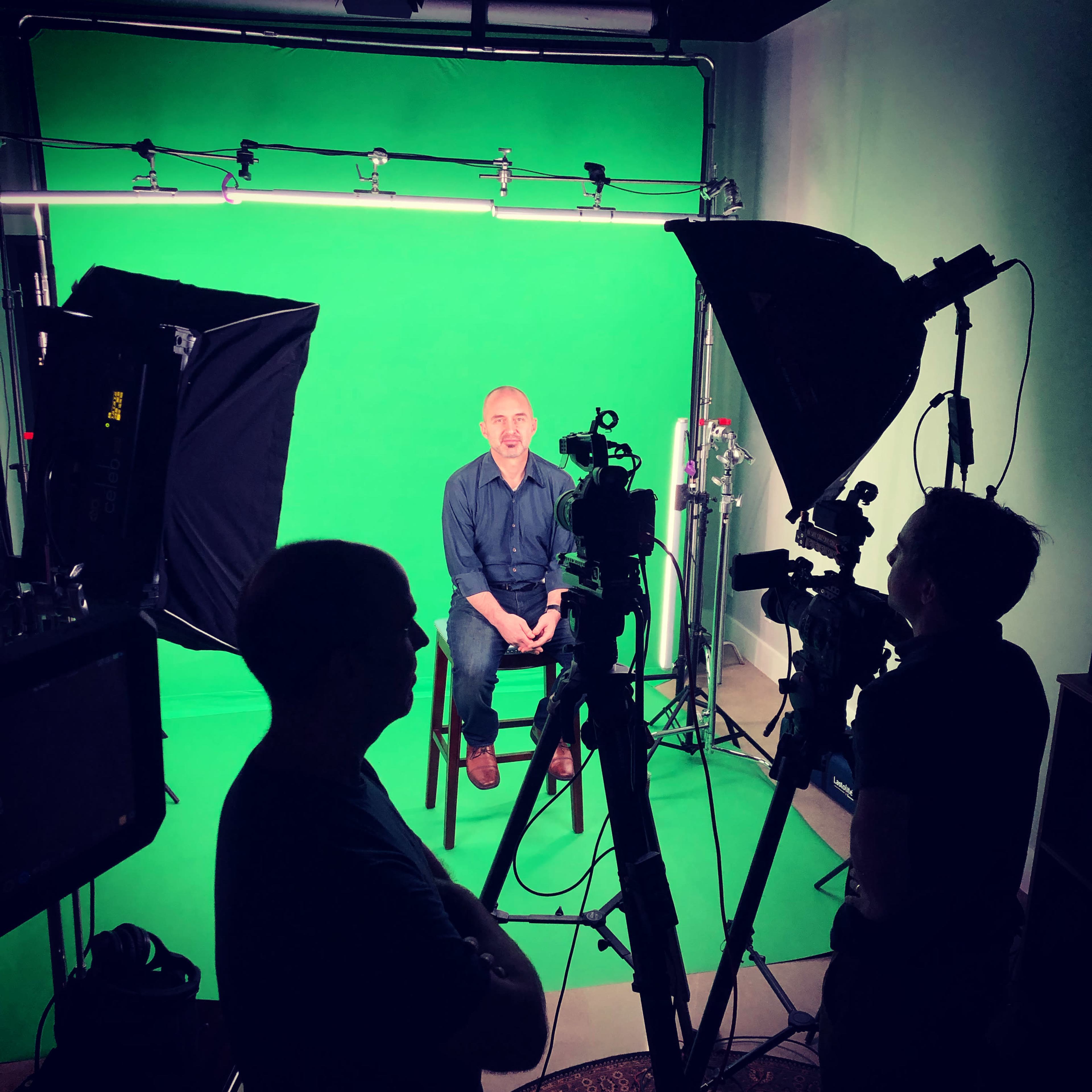 A person sits on a chair in front of a green screen, surrounded by multiple cameras and lighting equipment operated by two crew members.
