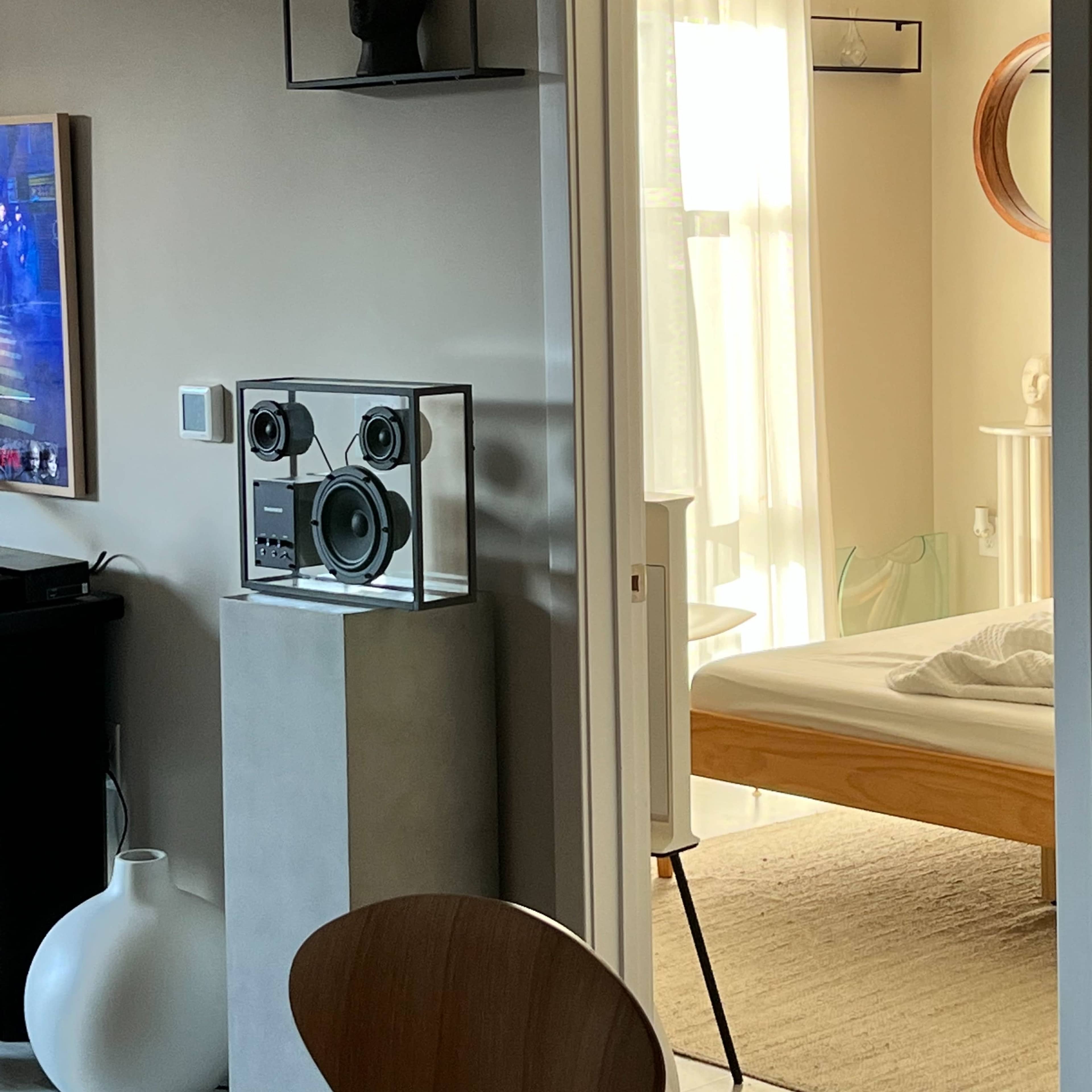A modern room with a console table holding a vintage speaker system, a wall-mounted bust display, and an open door leading to another room with a bed and natural light.