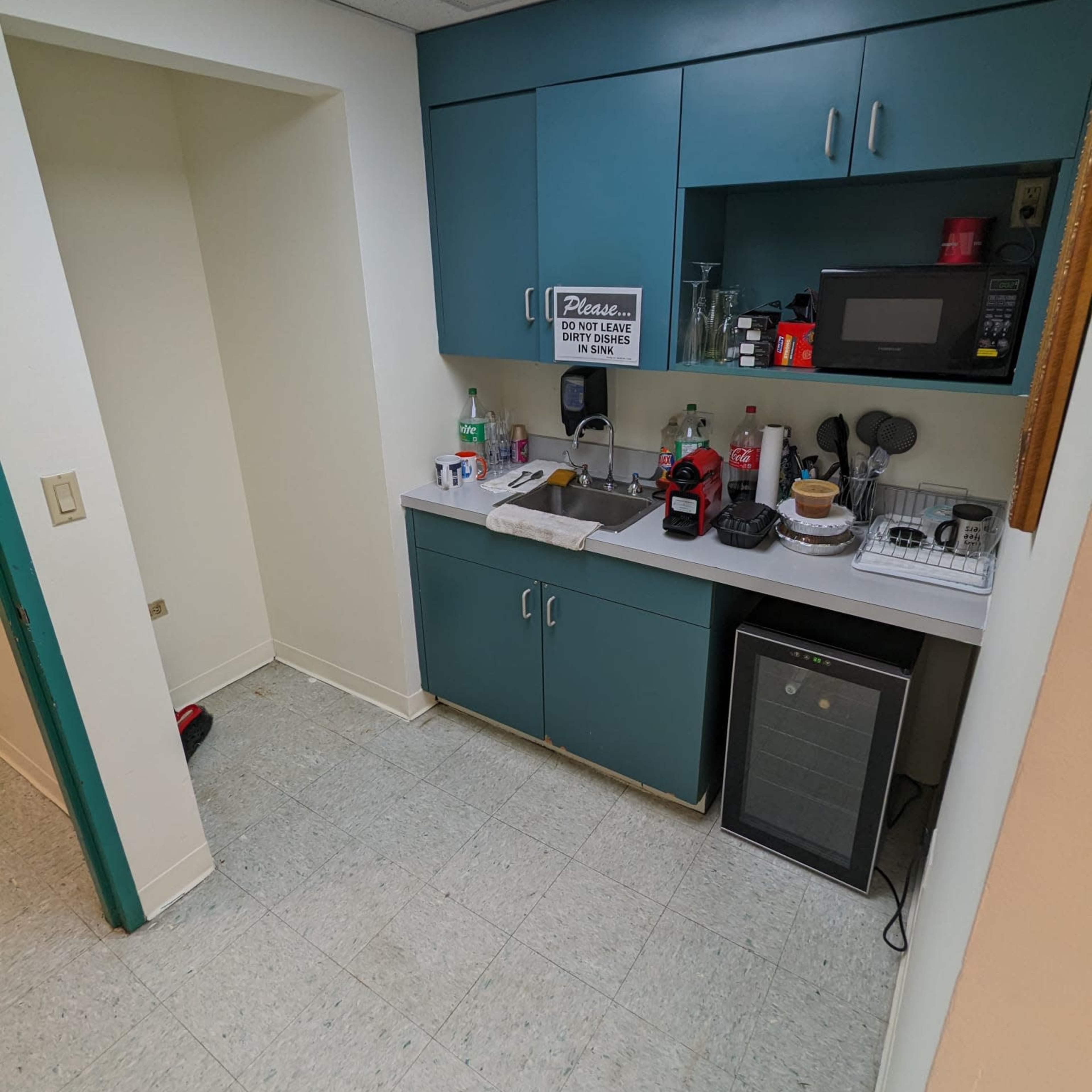 A small kitchen area with teal cabinetry, a microwave, and various kitchen items on the counter, including bottles and utensils.