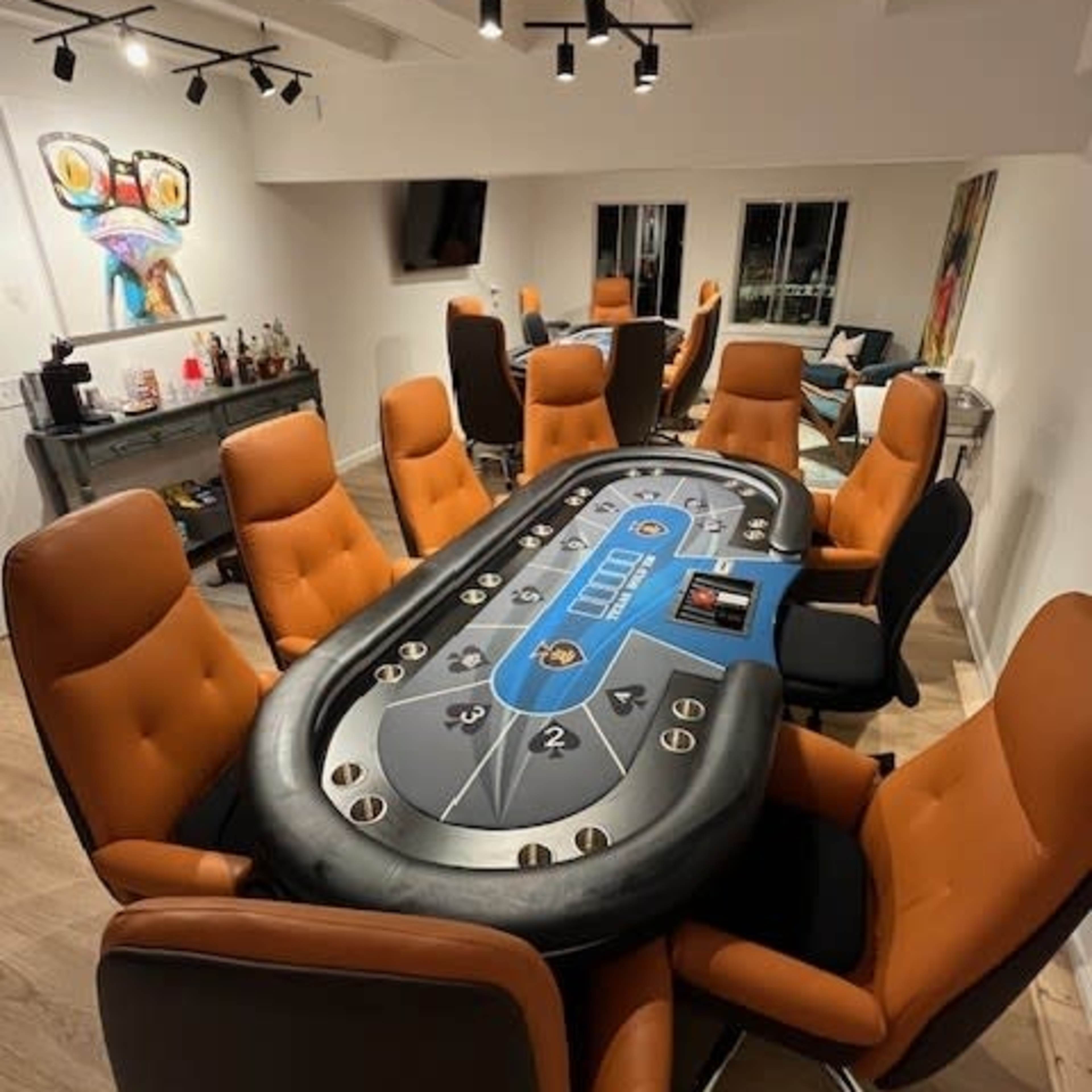 A modern poker room with a large oval table surrounded by orange upholstered chairs and a bar featuring several bottles in the background.