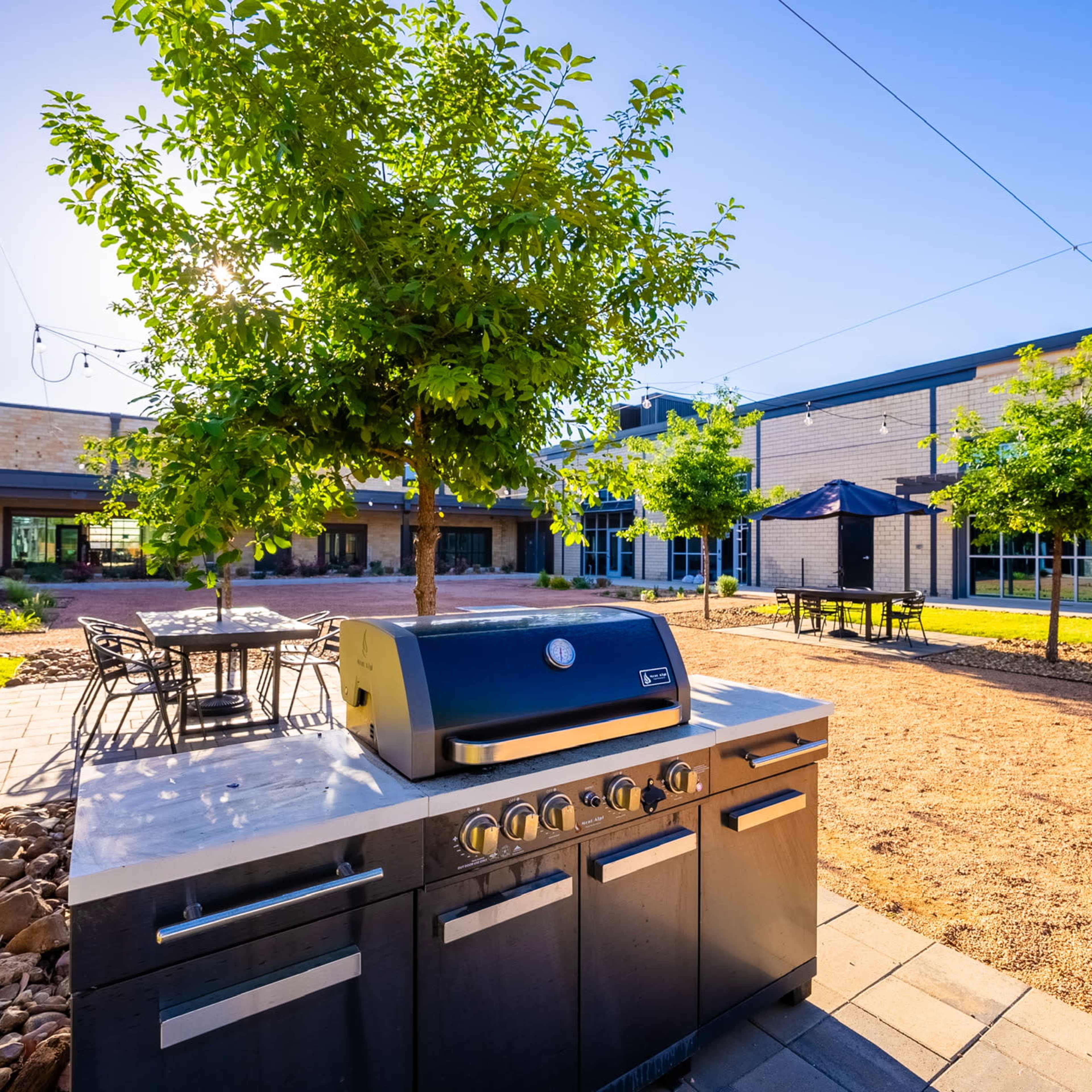 The scene features an outdoor grill station surrounded by a landscaped courtyard with trees, picnic tables, and stone pathways.