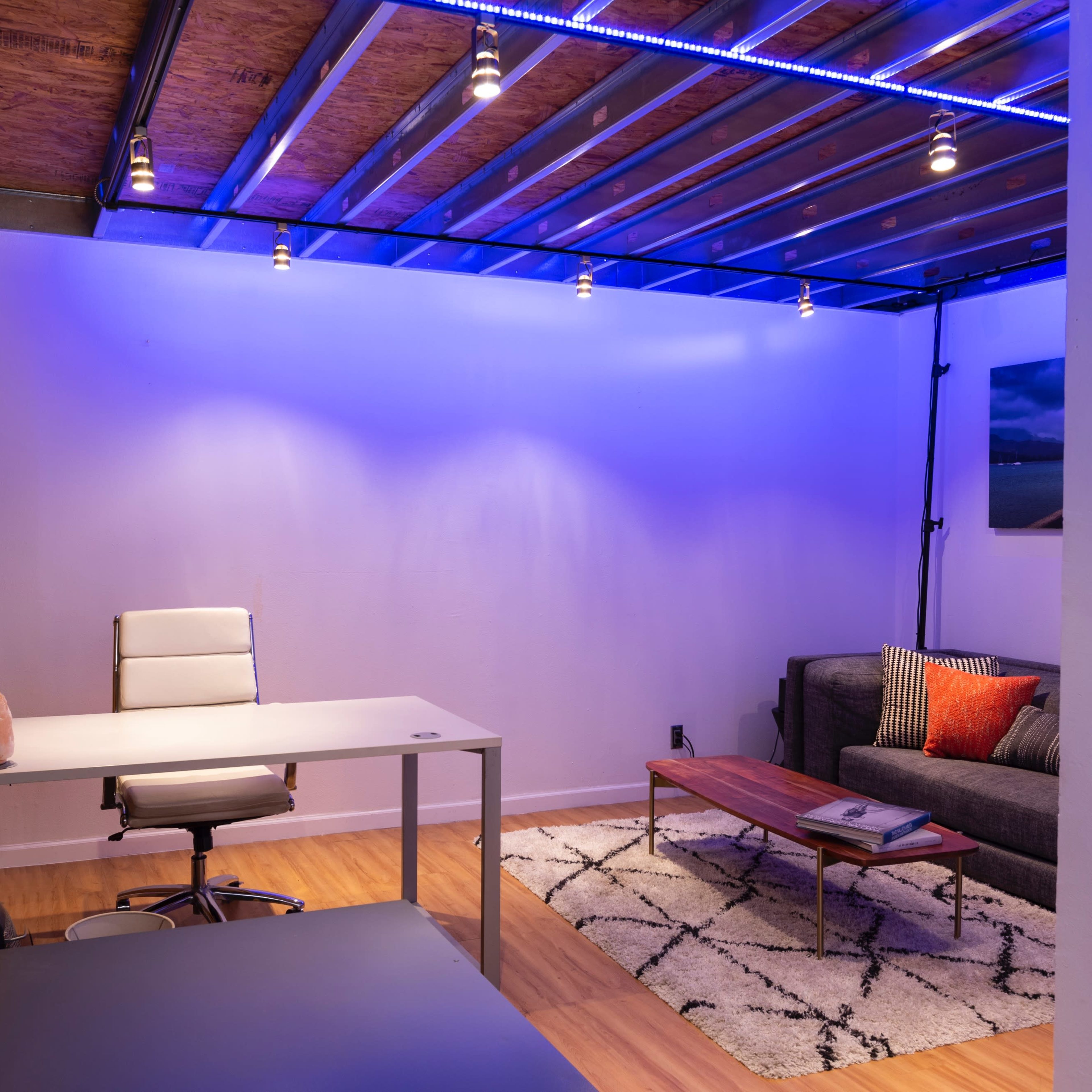 A modern office space featuring a desk and chair, a sofa, a coffee table, and ambient lighting along the ceiling.