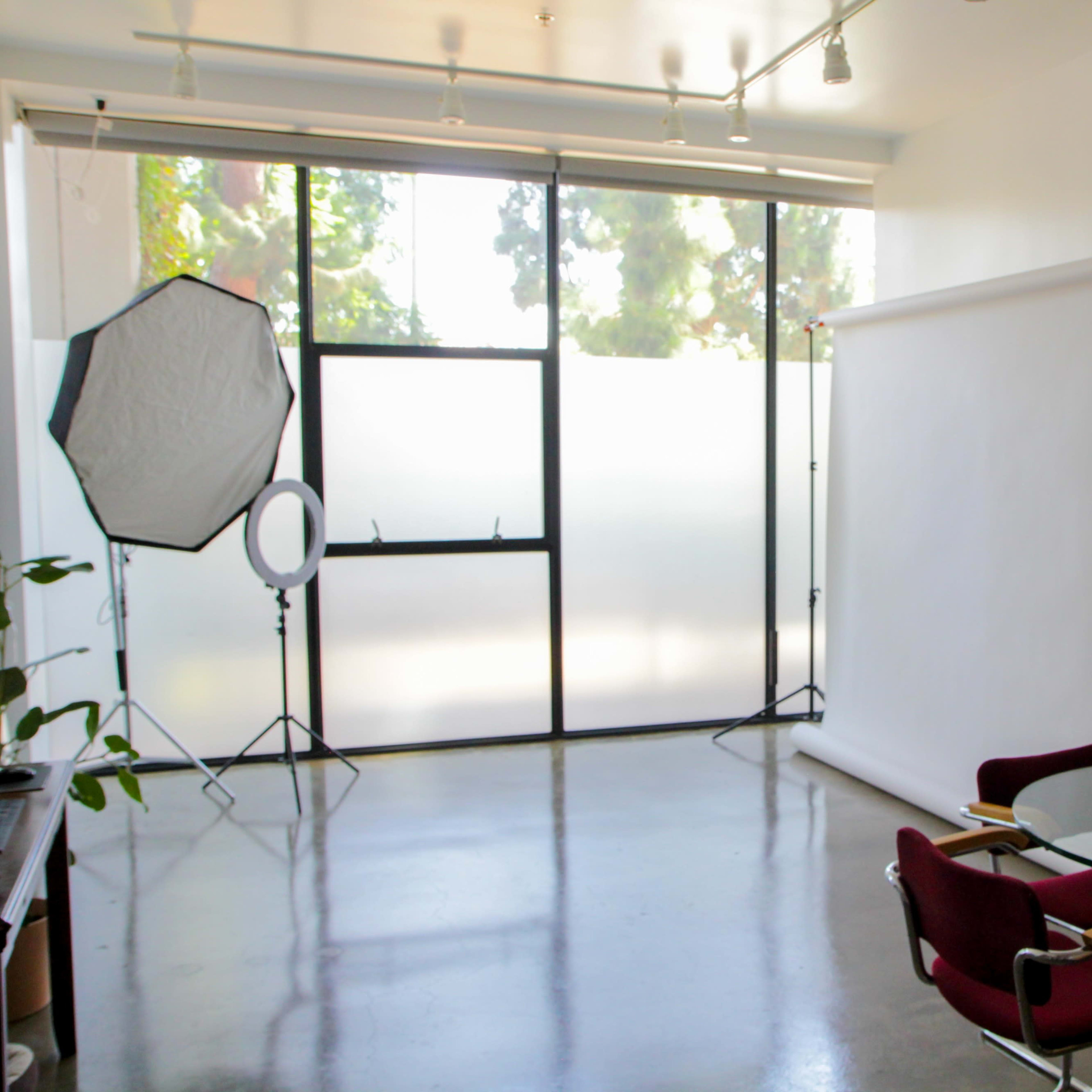 A bright, minimal studio space with large windows, a backdrop setup, and a glass table surrounded by red chairs.