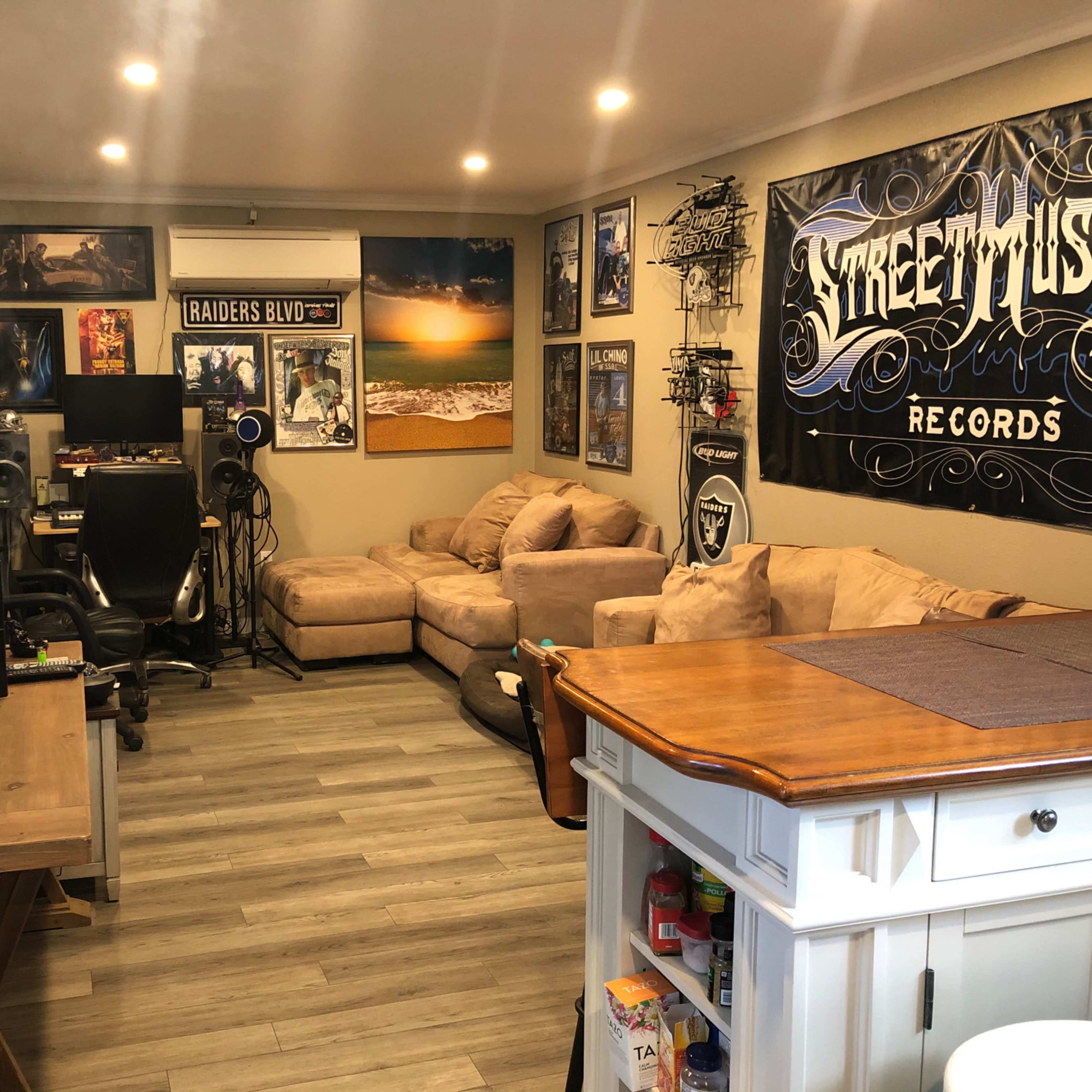 A room with beige couches, a small dining table, various framed posters on the walls, and audio equipment set up in the corner.