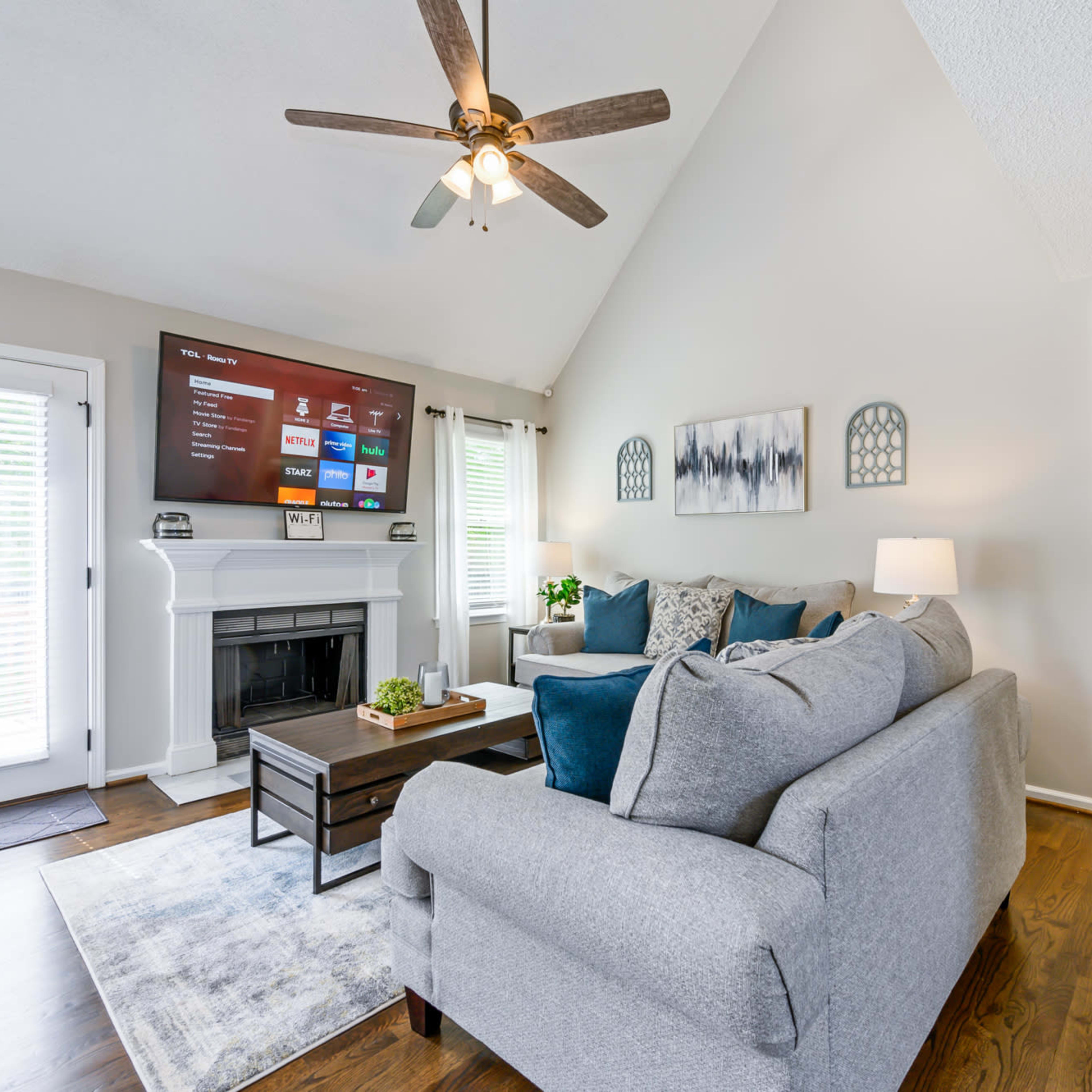 A living room features a gray sectional sofa, a wooden coffee table, and a mounted television above a fireplace, all illuminated by natural light.