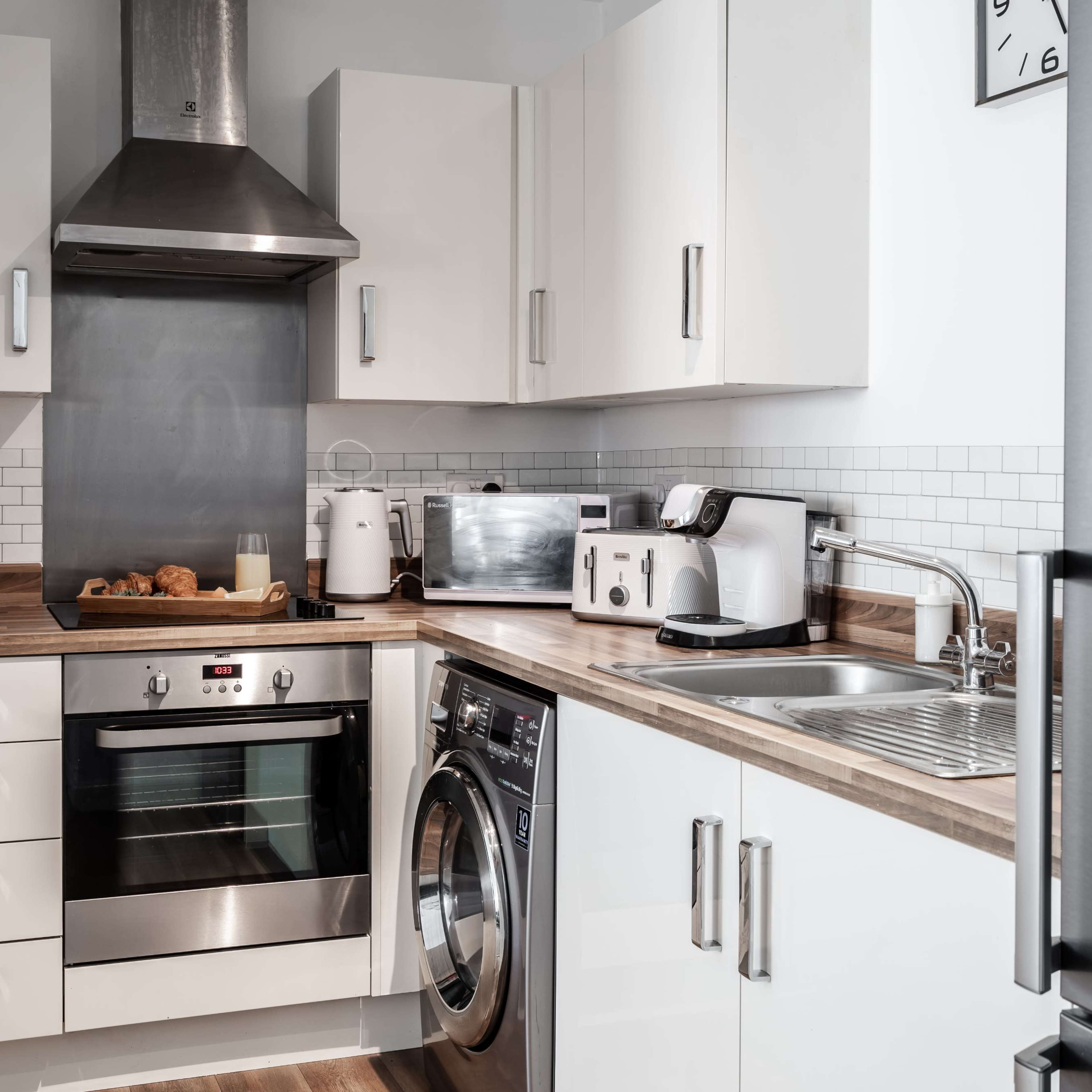 A modern kitchen featuring an oven, washing machine, and various appliances against a light-colored backdrop.