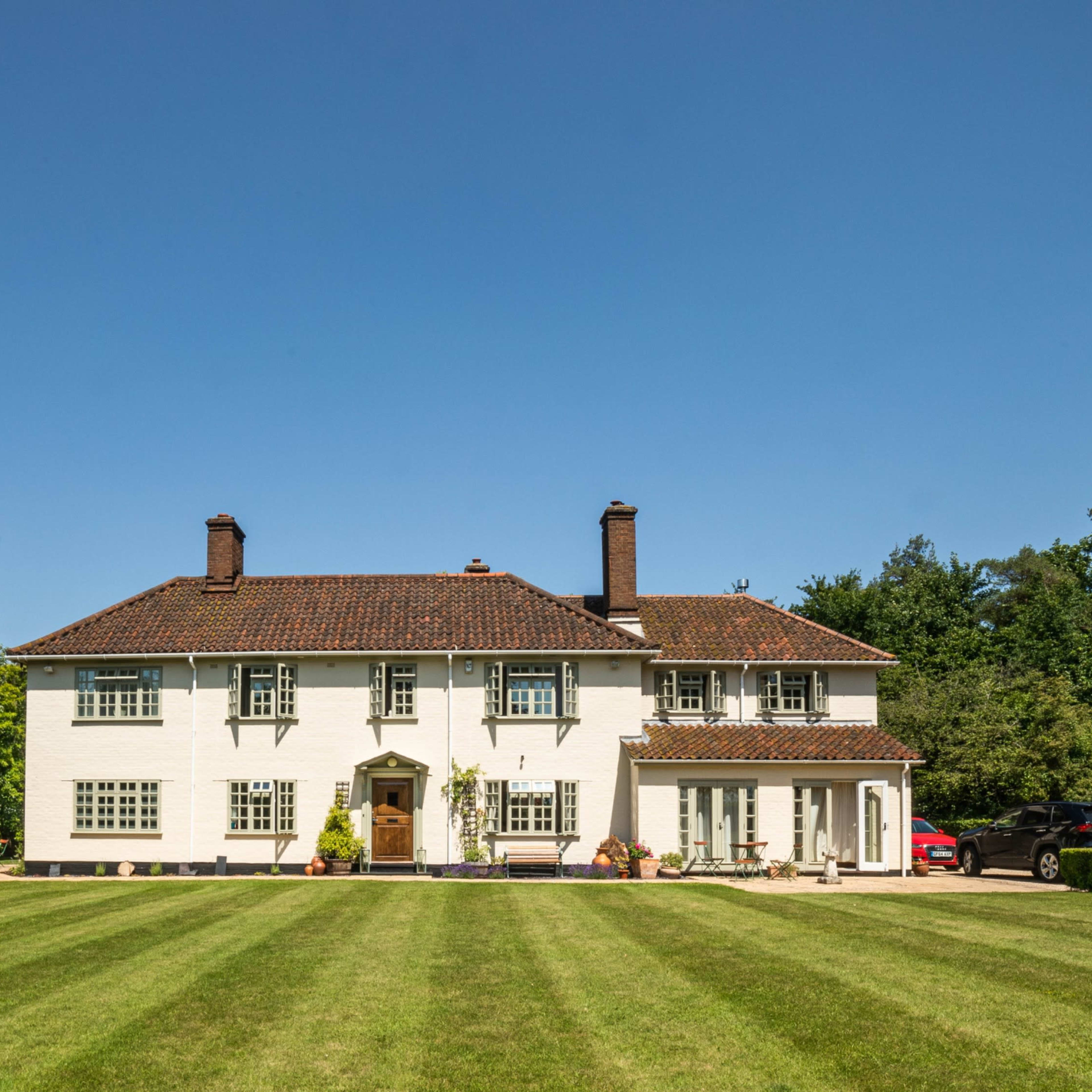 A two-story white house with a tiled roof sits on a well-maintained lawn surrounded by trees.