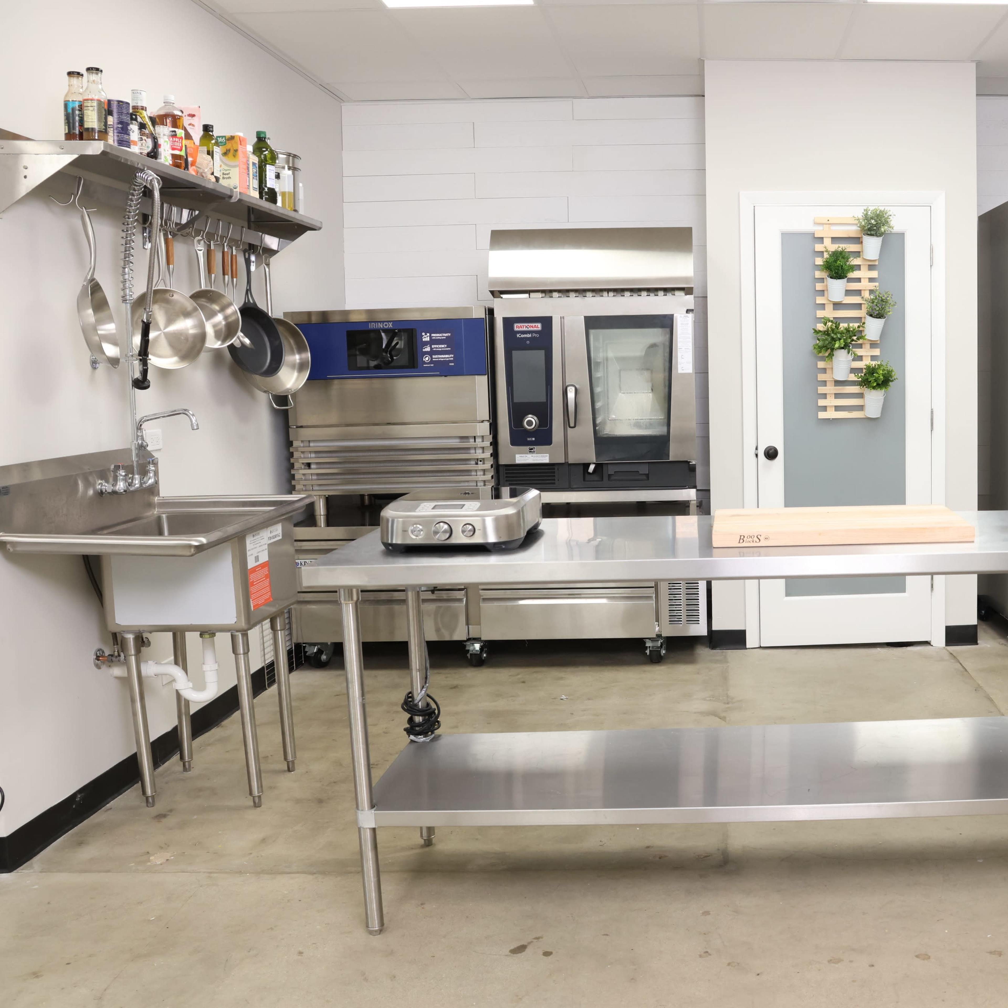 A modern commercial kitchen with stainless steel countertops, a sink, and various kitchen appliances including a refrigerator and an oven.