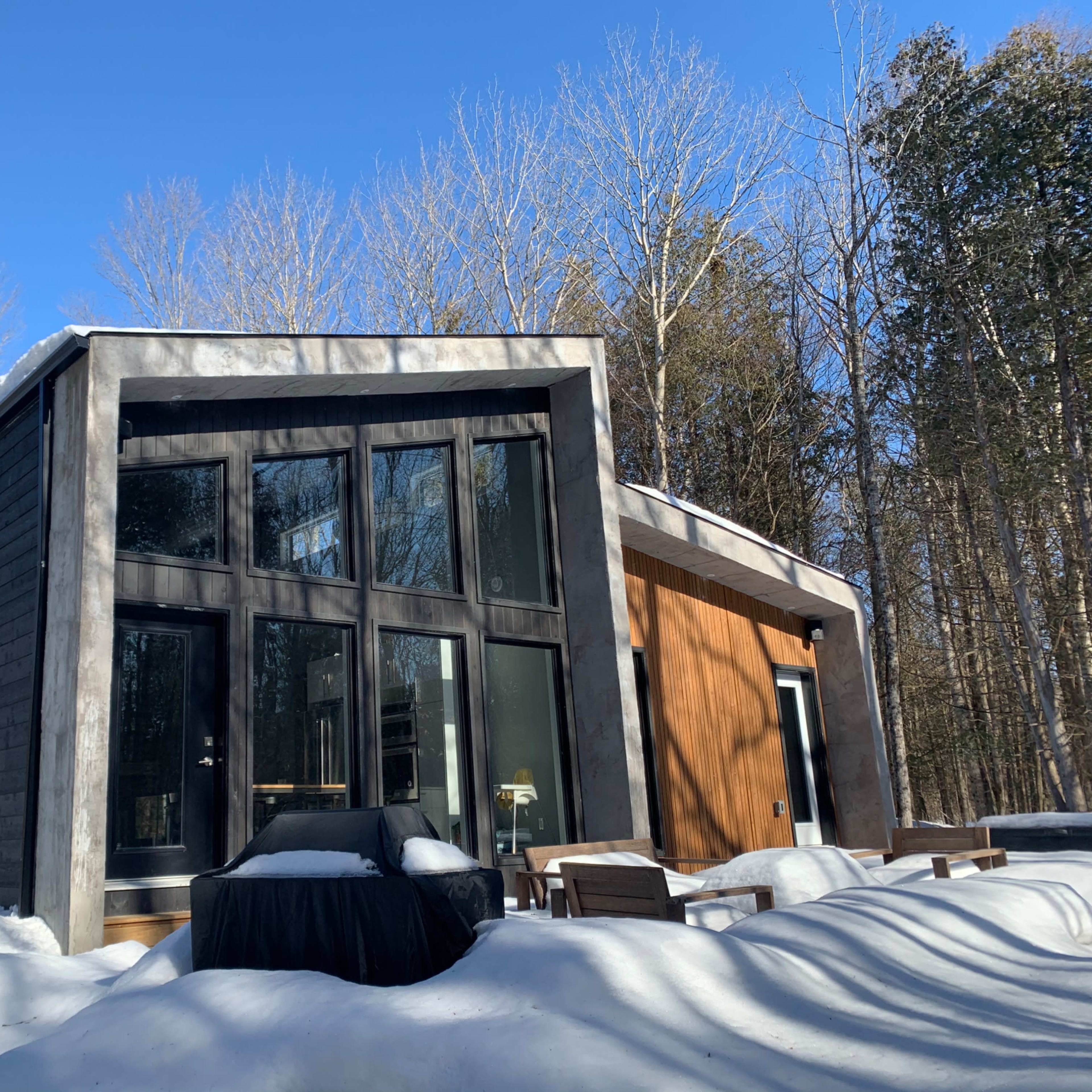 A modern building with large windows and a wooden facade is partially covered by snow, surrounded by bare trees under a clear blue sky.