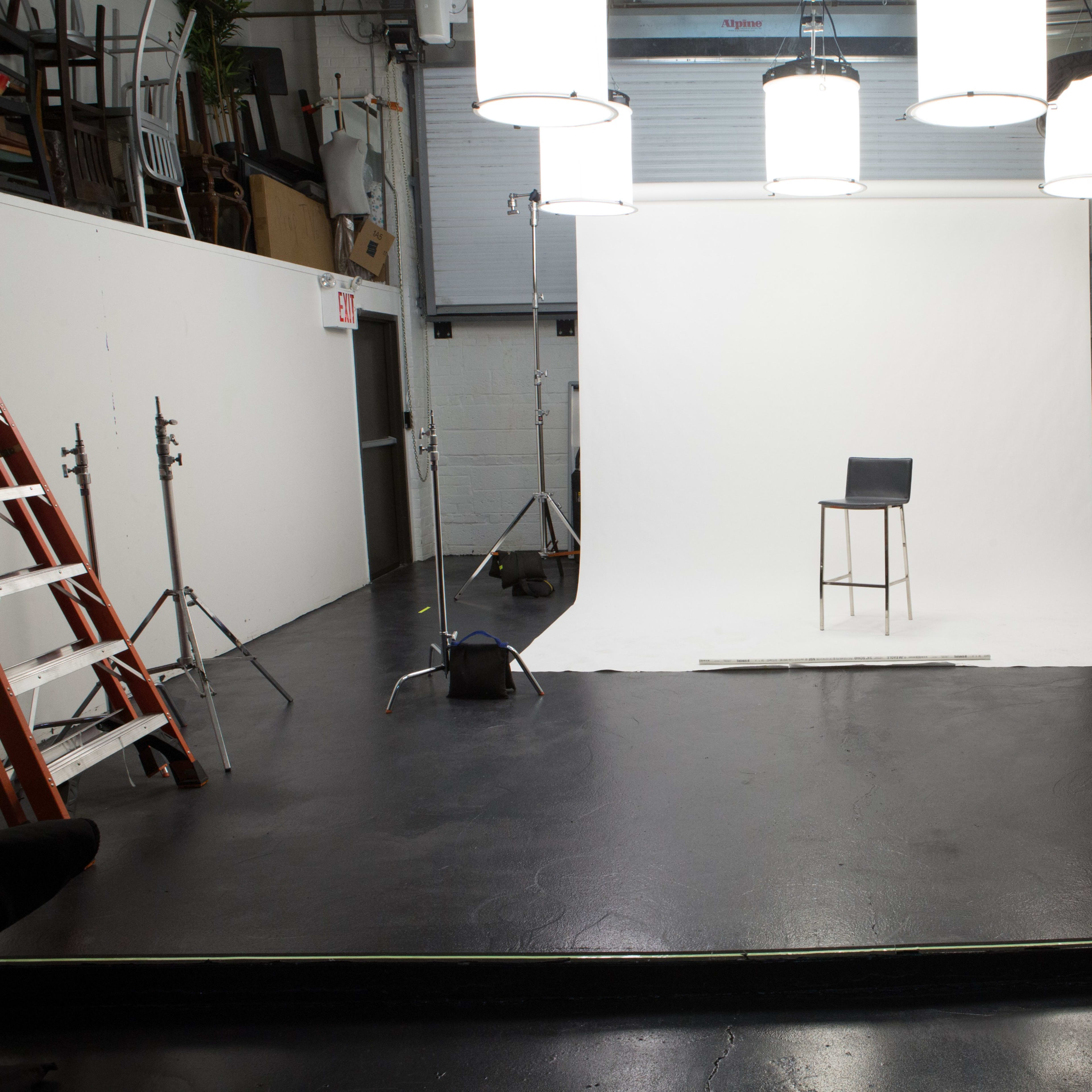 A photography studio with a white backdrop, a tall stool in the center, and several light fixtures hanging above, along with equipment and ladders positioned around the space.