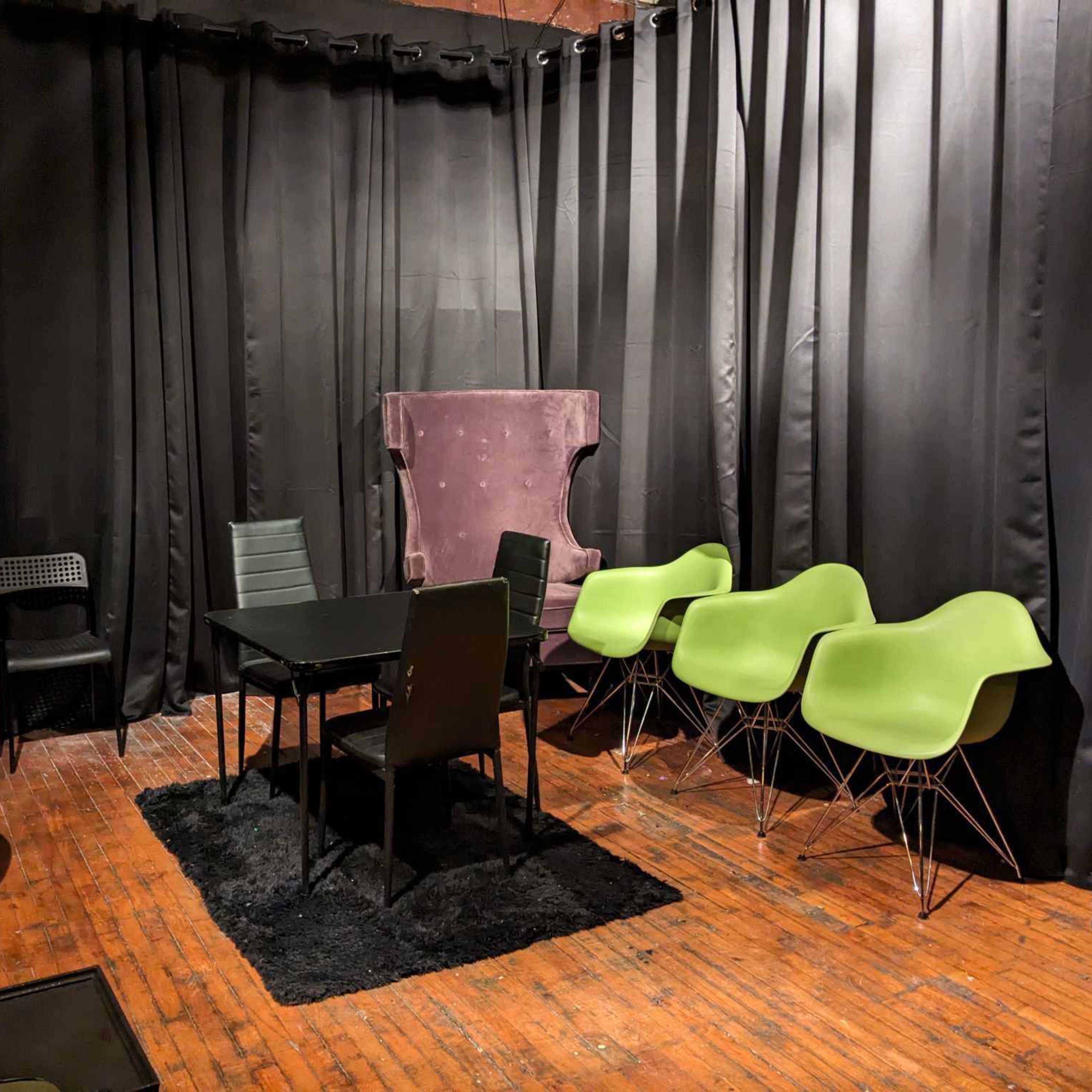A room with black curtains, a round table surrounded by various chairs, and a large purple armchair in the corner.