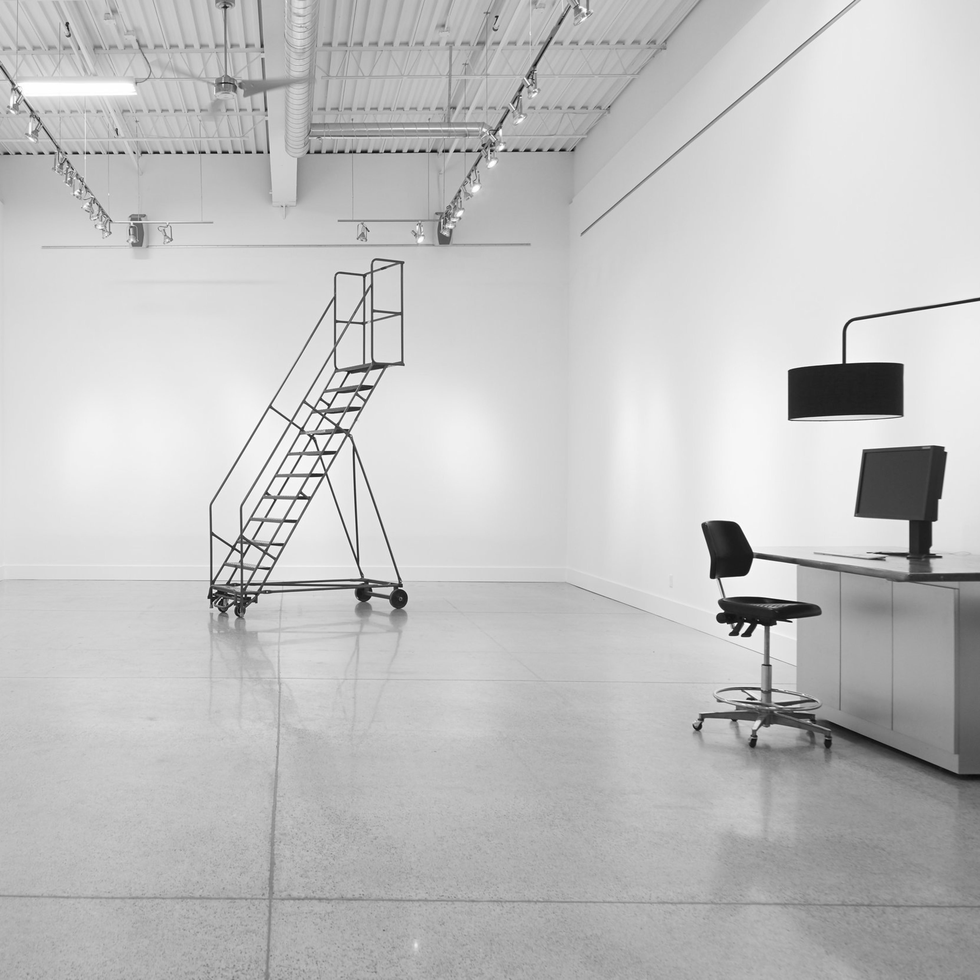 A spacious, minimalistic room with a metal ladder, a desk with a chair, and a computer setup against a stark white wall.