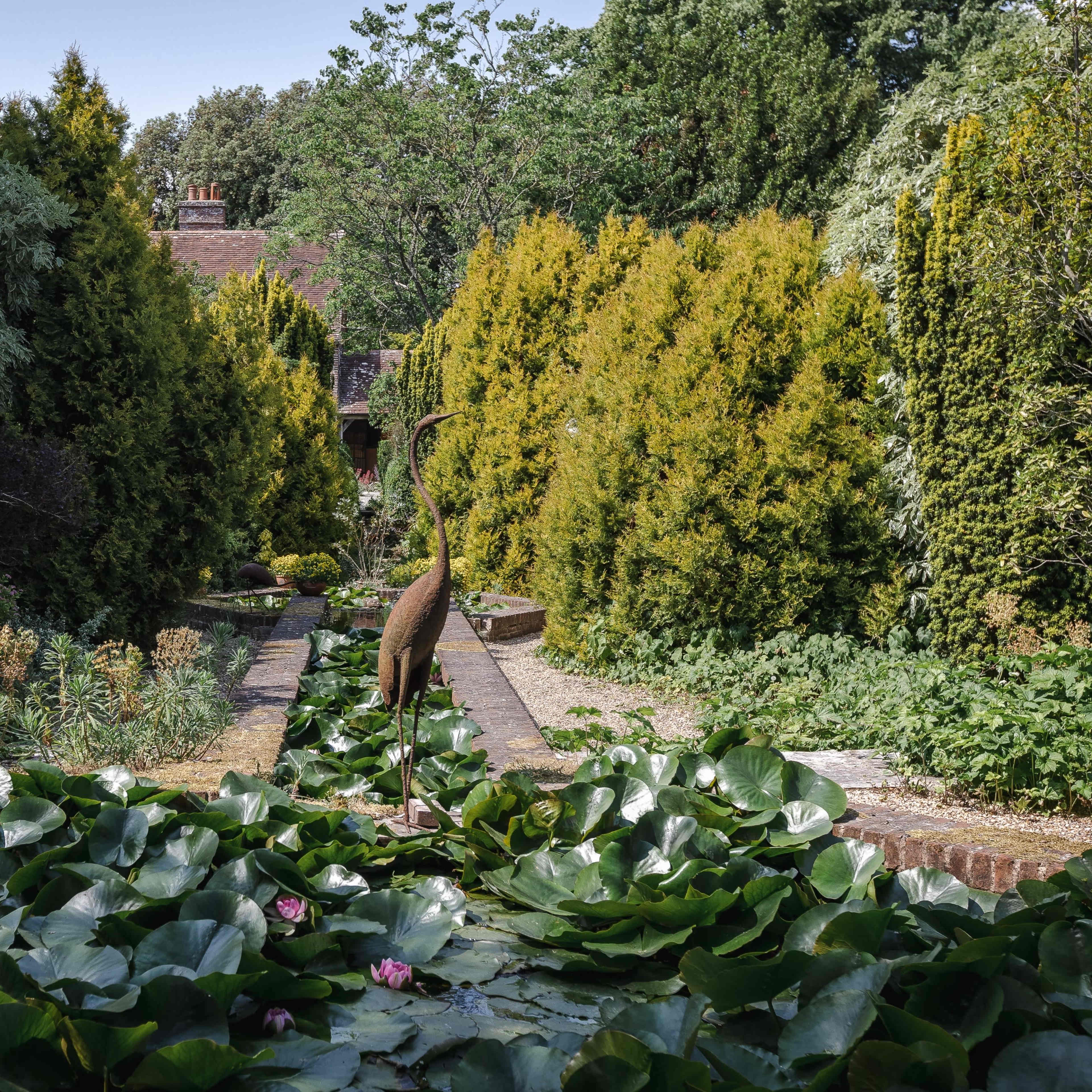 A metal sculpture of a heron stands amidst a garden filled with large lily pads and lush greenery.