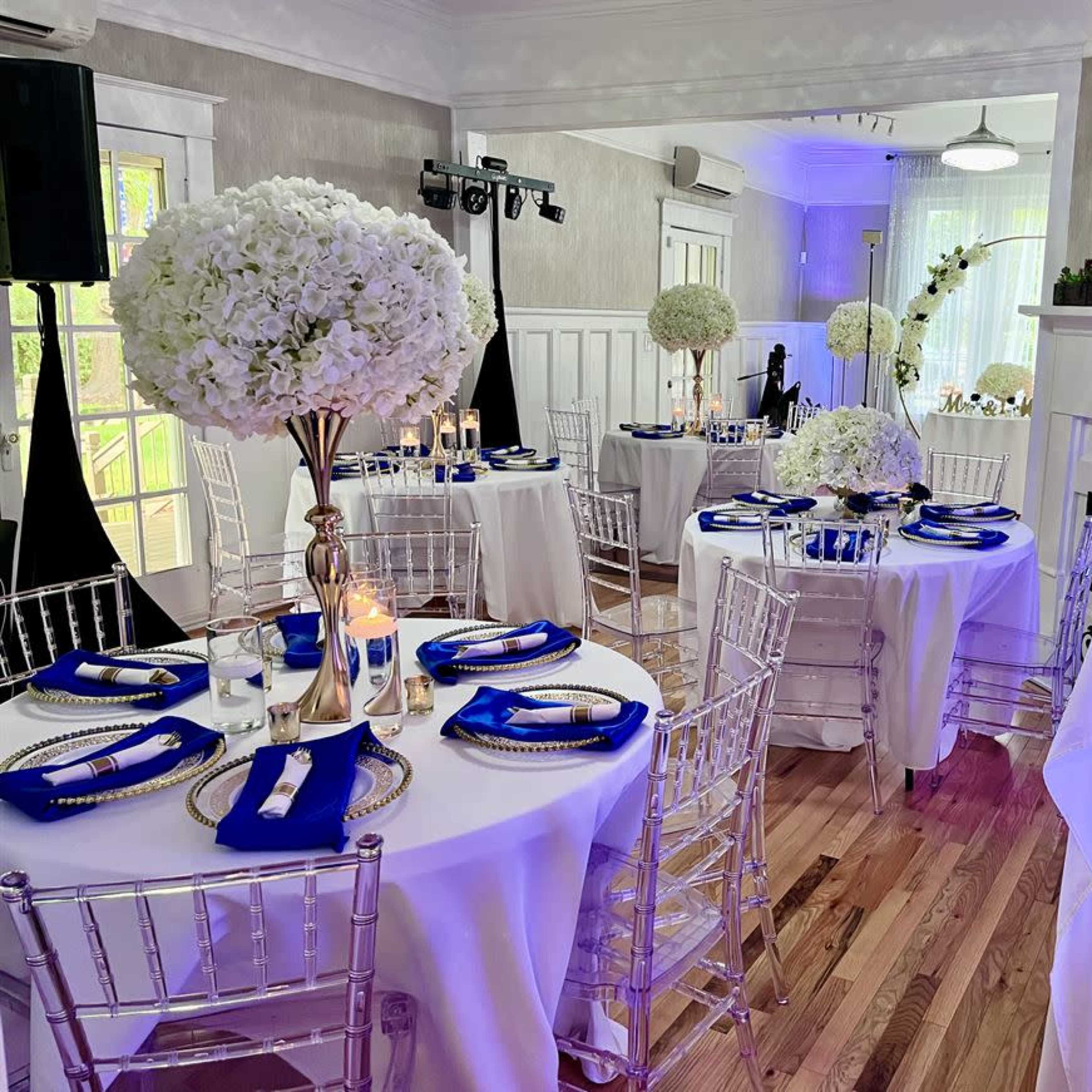 A decorated event space with round tables covered in white tablecloths, each featuring a tall centerpiece of white flowers and blue napkins, set up for a formal gathering.