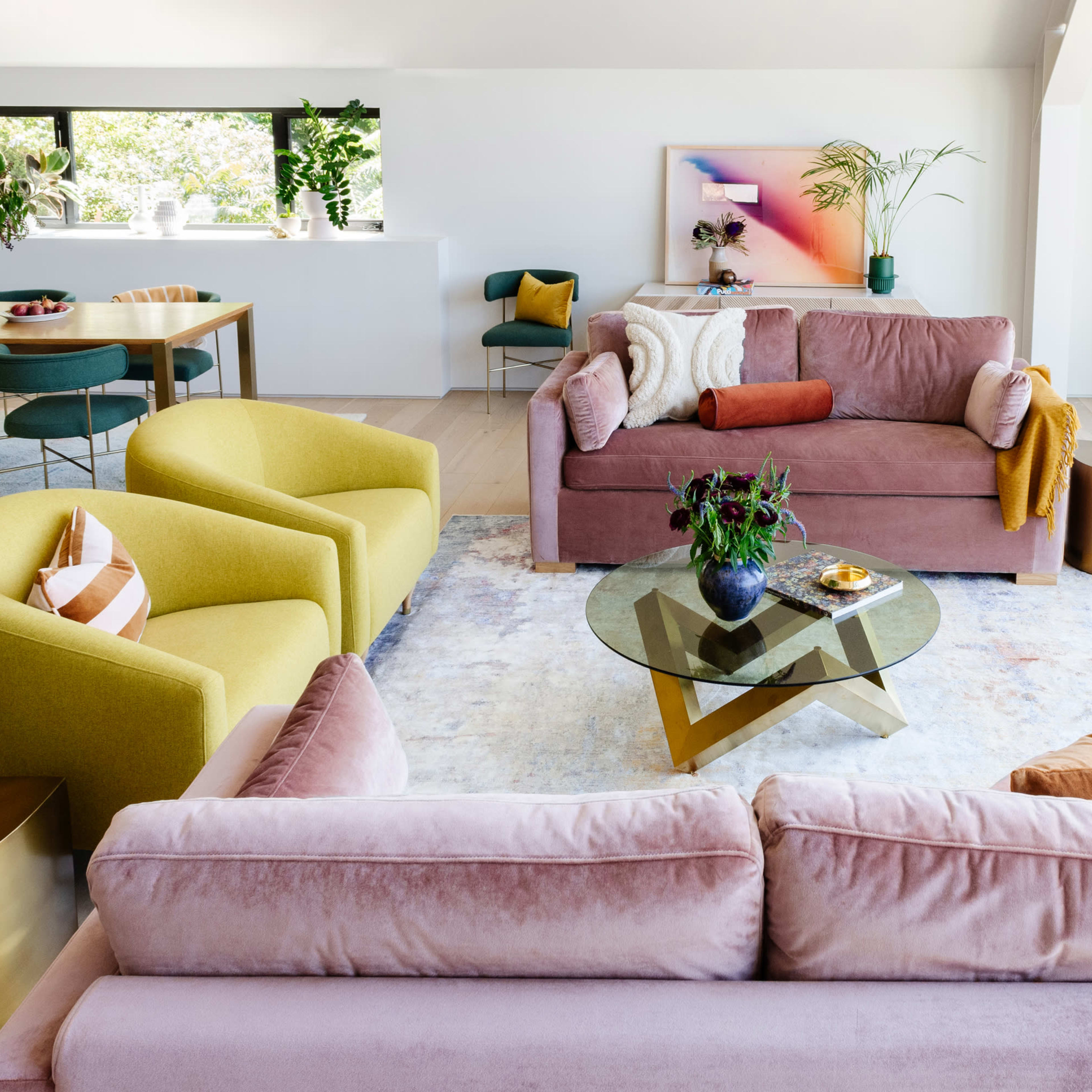 A modern living room features two couches, colorful armchairs, a glass coffee table, and large windows letting in natural light.