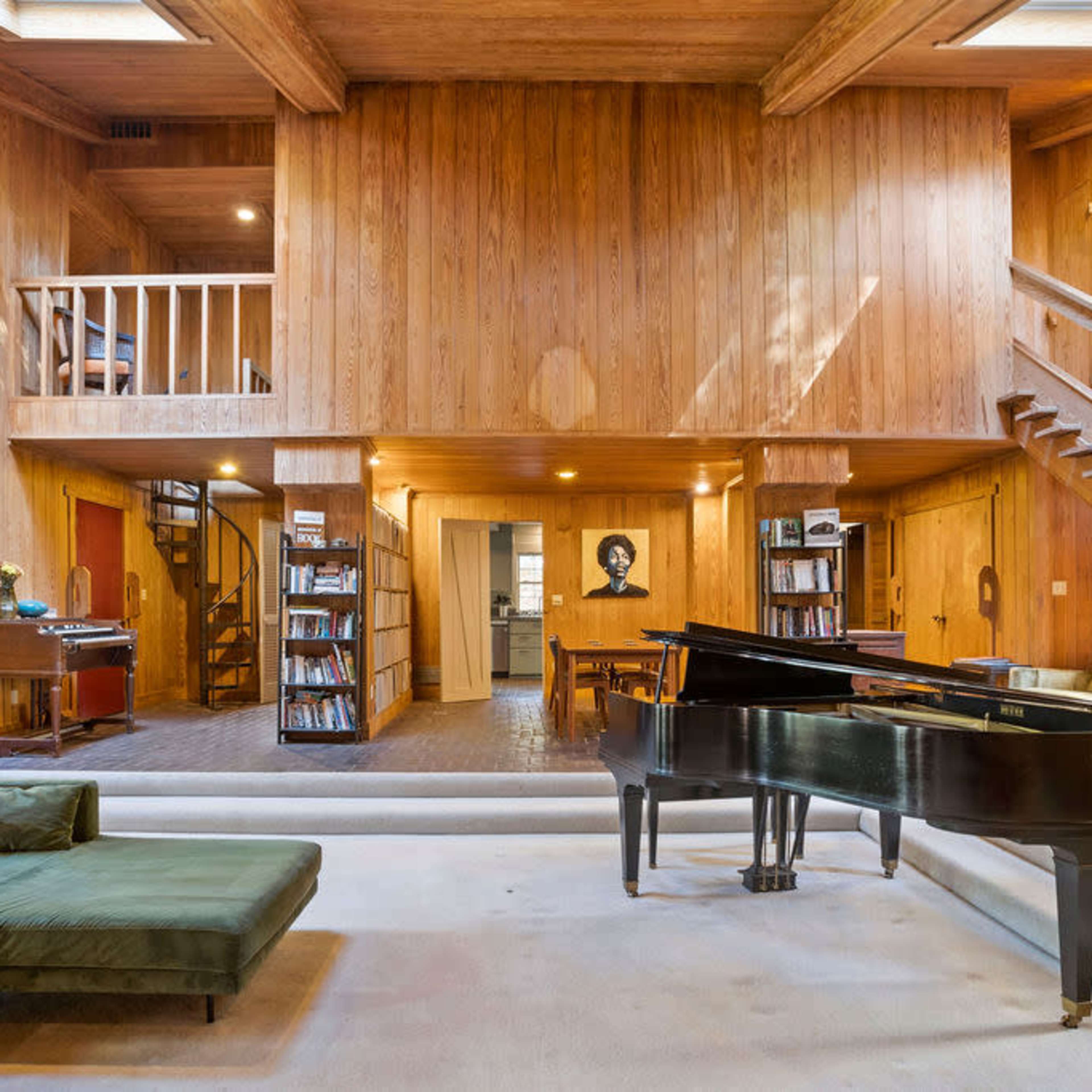 A spacious, wooden-paneled living area featuring a grand piano, a staircase, and a second-floor balcony overlooking the main space.