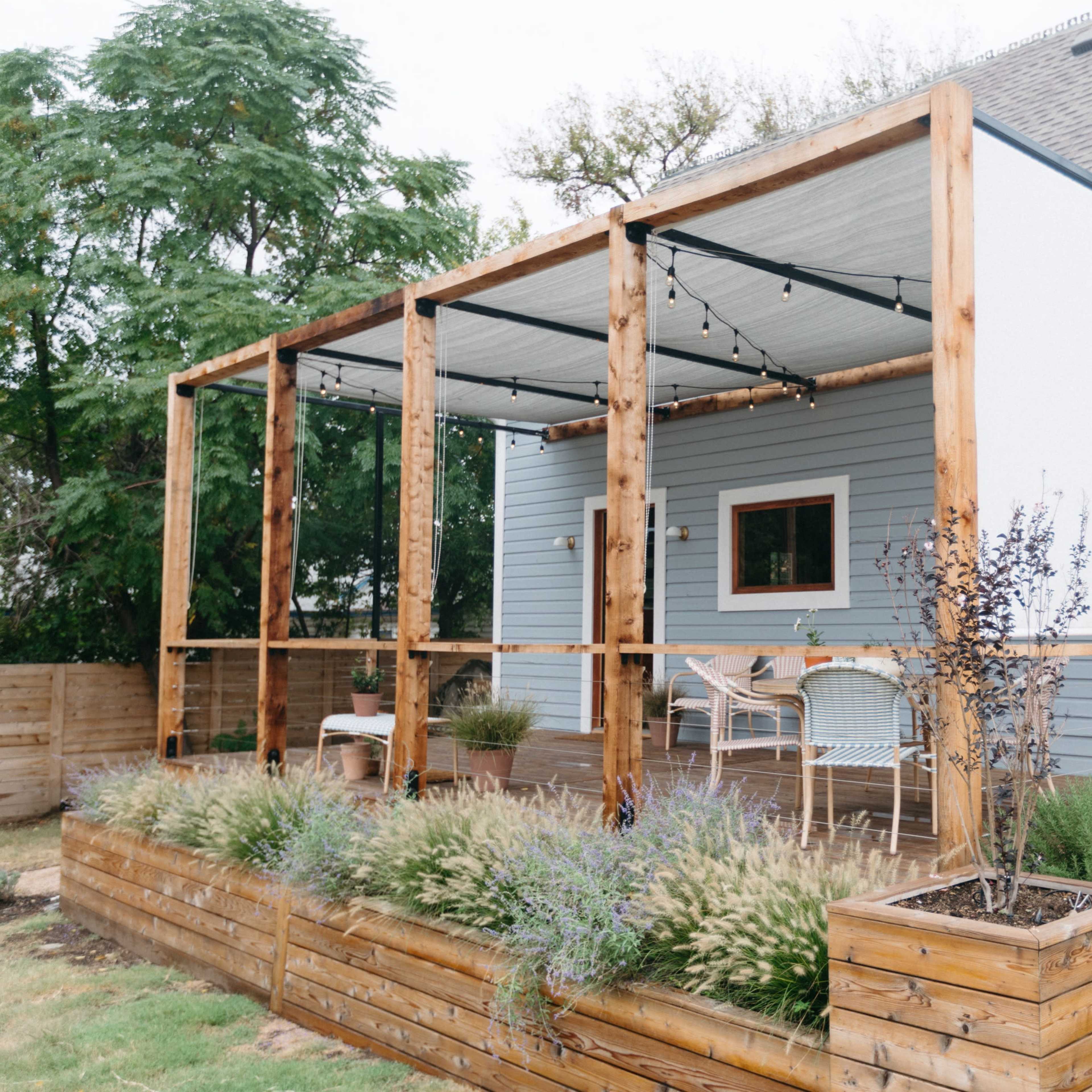 A modern wooden patio structure attached to a gray house, featuring a covered seating area and garden beds with plants.