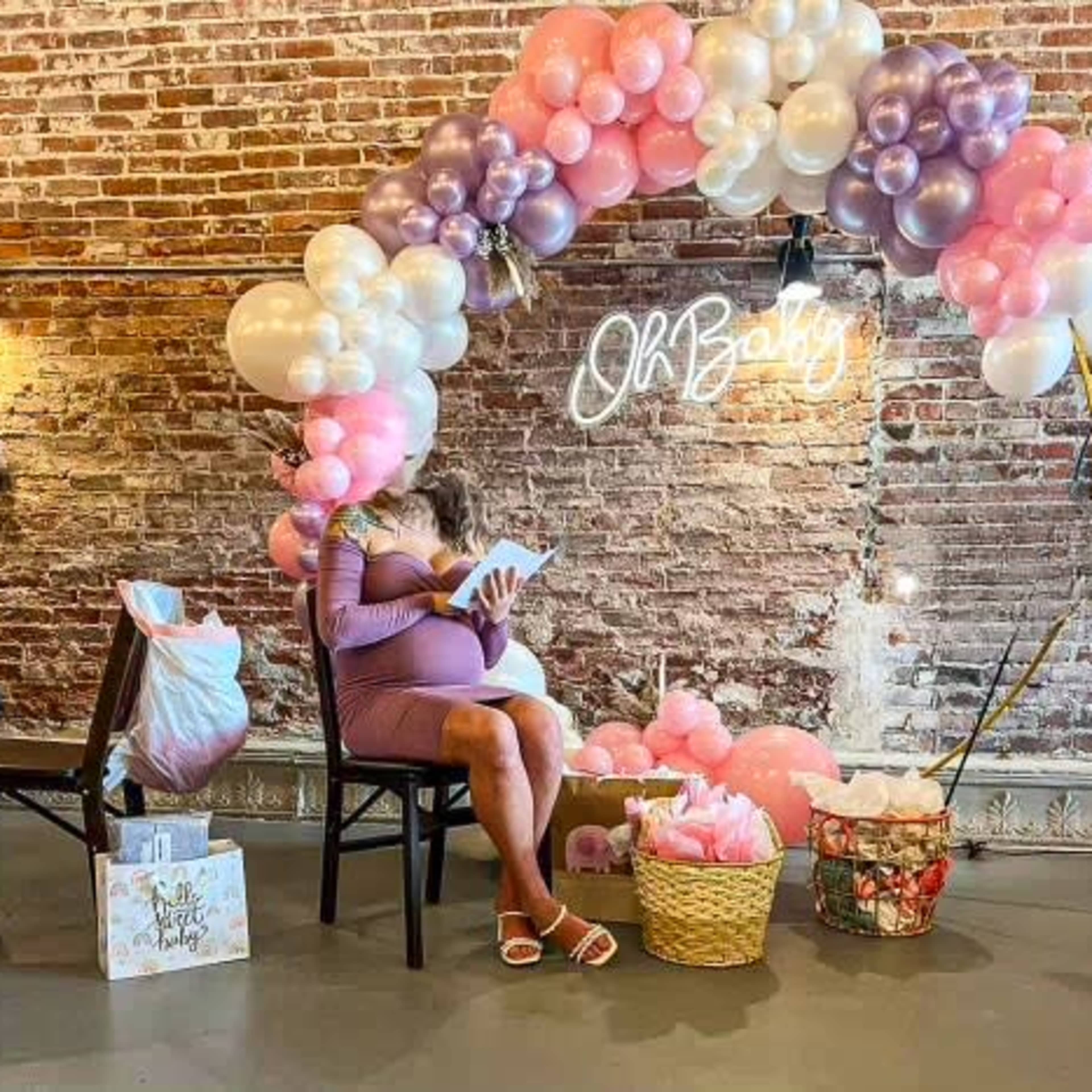 A pregnant woman sits in a chair under a balloon arch while reading a book in a decorated space featuring gifts and decorations for a baby shower.