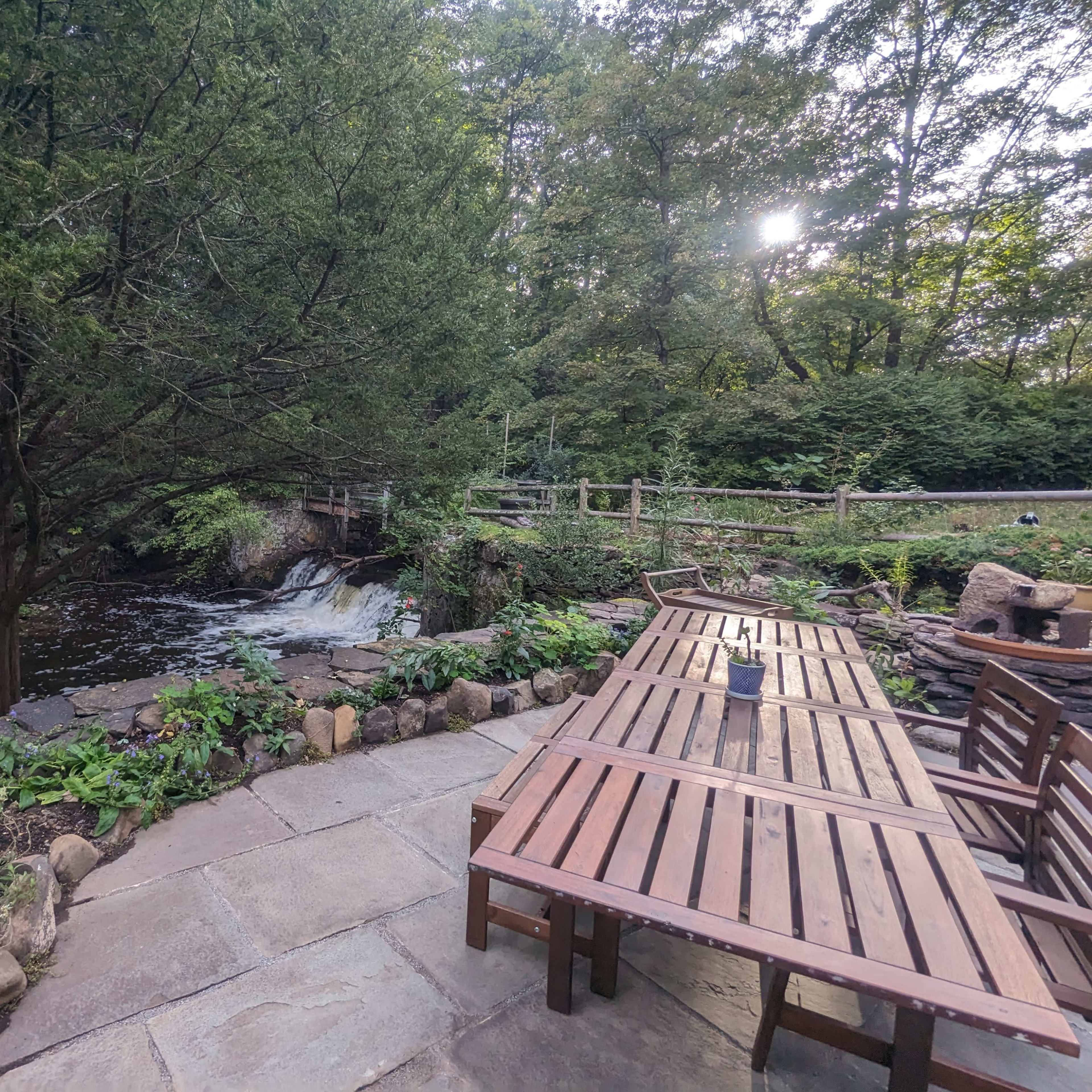 A wooden table and benches are set along a stone path near a flowing stream surrounded by trees.