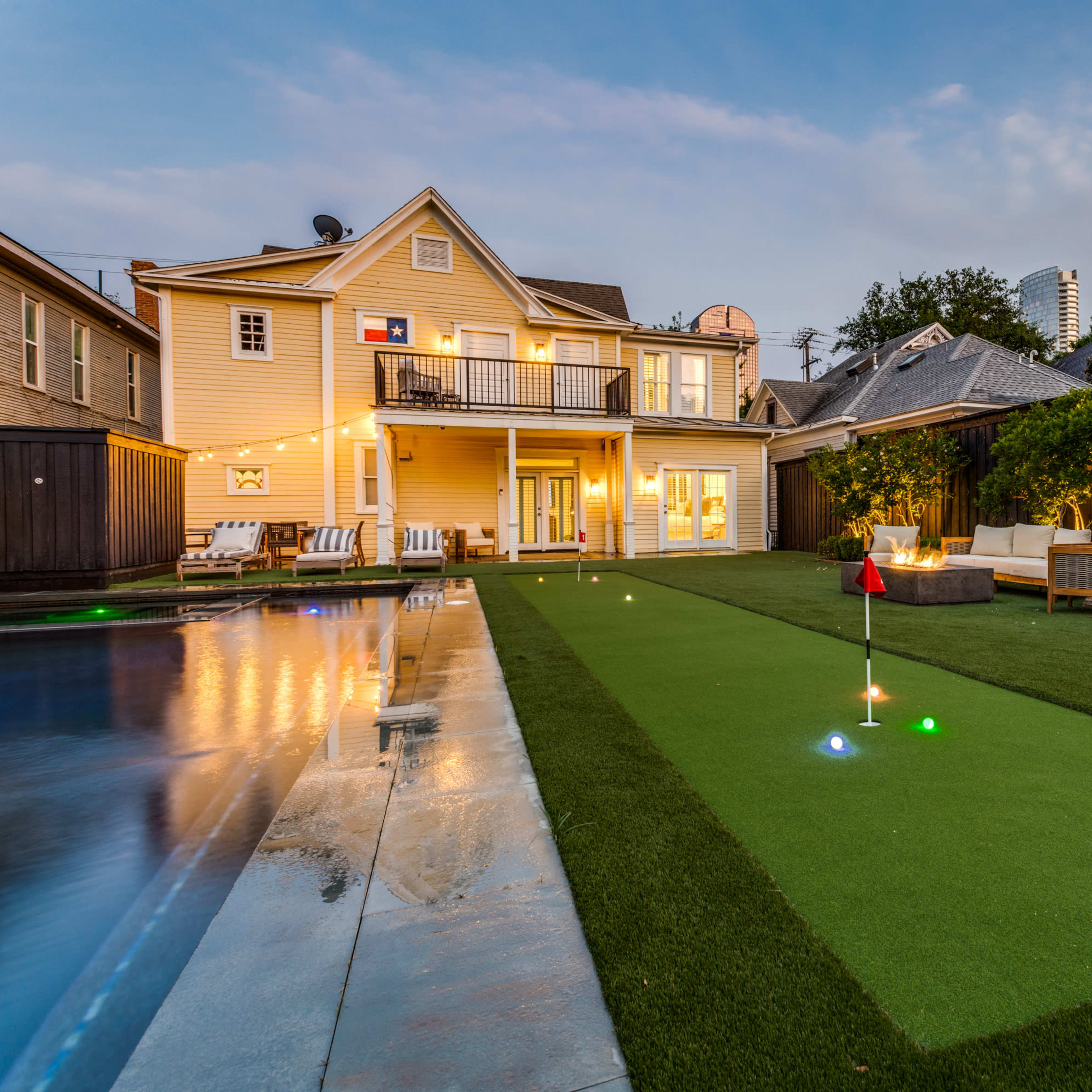 A two-story yellow house features a well-maintained backyard with a putting green and a swimming pool, illuminated by landscaping lights at dusk.