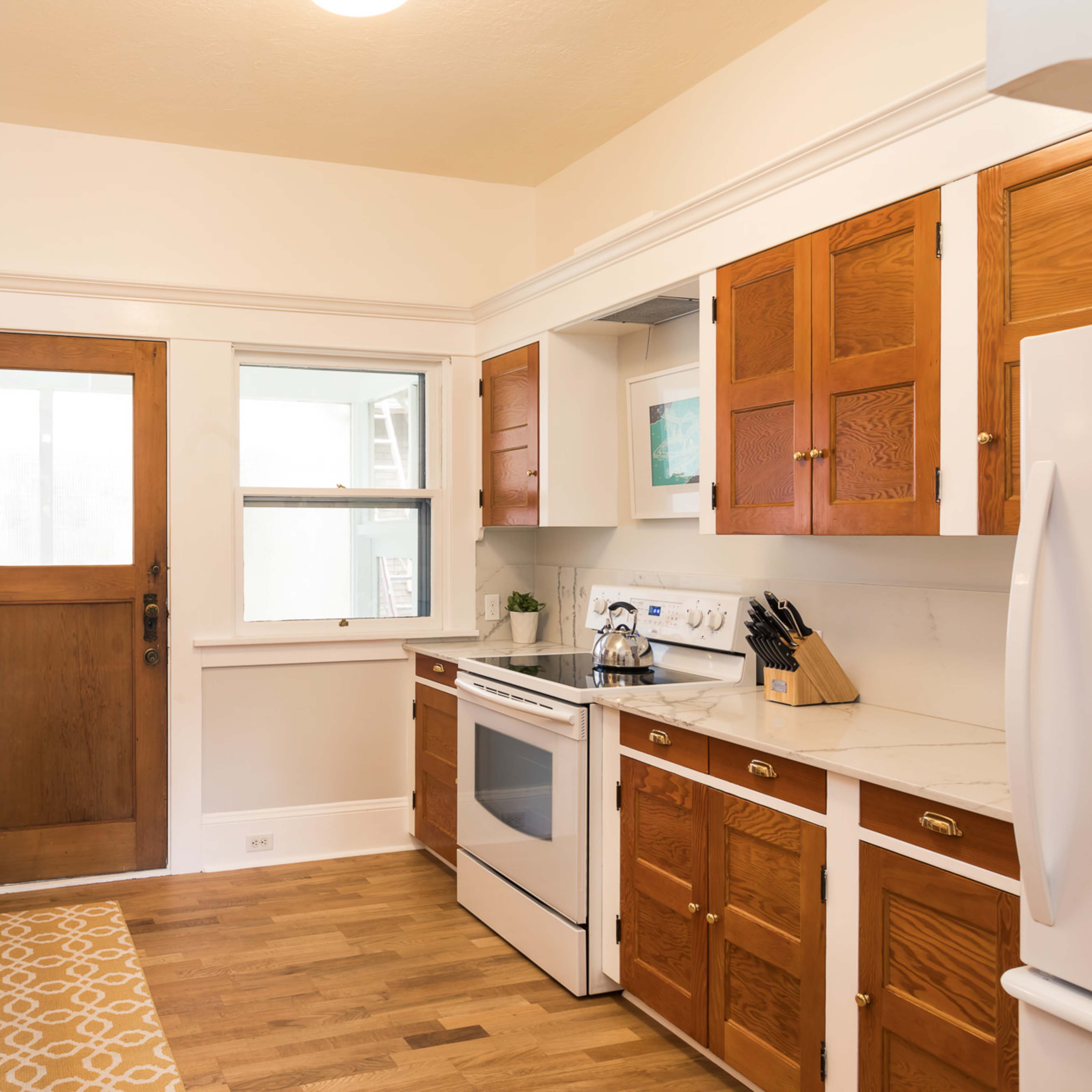 A kitchen features white walls, wooden cabinetry, and a marble countertop with a window and door leading outside.