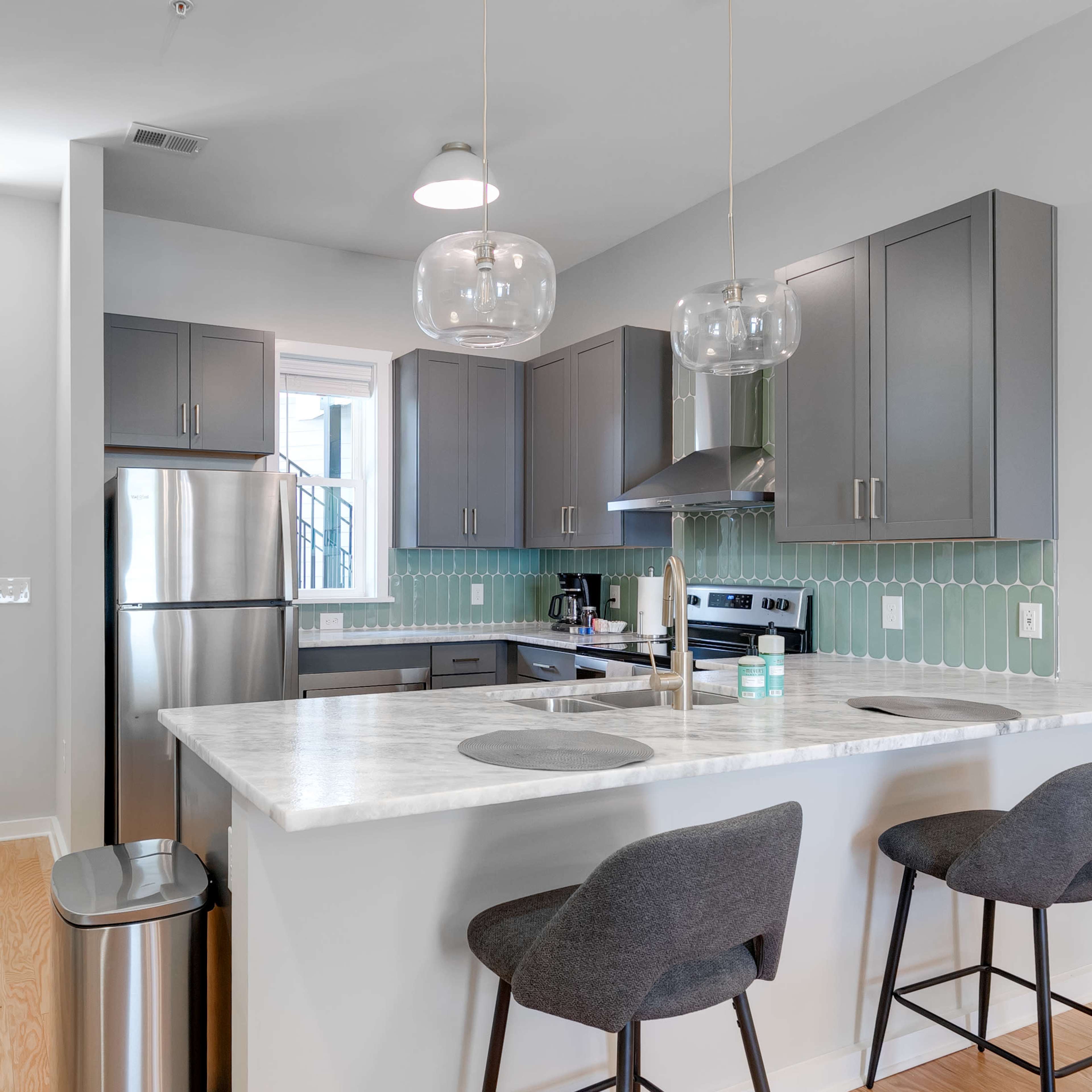 A modern kitchen with gray cabinets, a marble countertop, and two bar stools, connected to a living area with a door leading outside.
