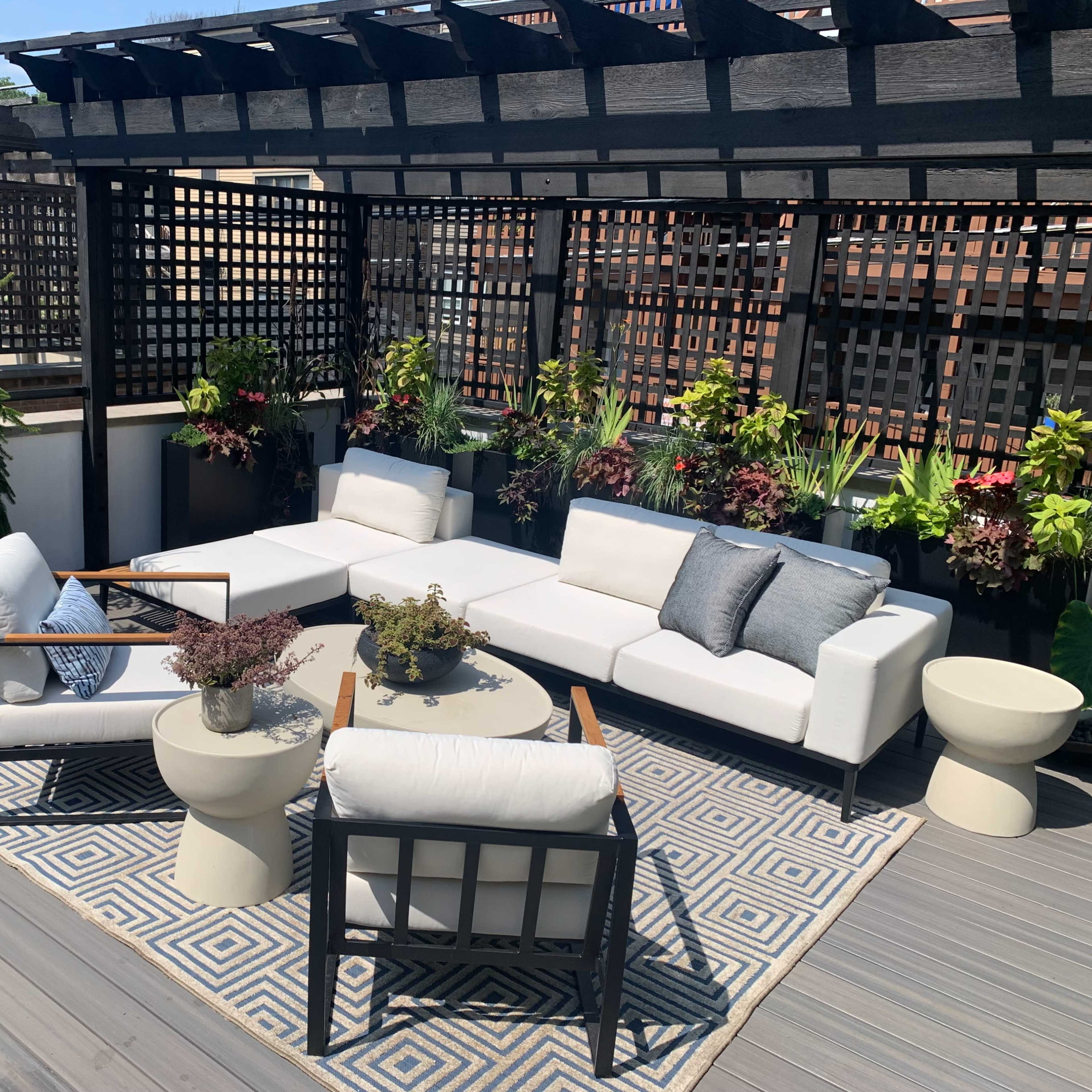A rooftop patio featuring modern seating arrangements, potted plants, and a geometric rug under a trellis structure.