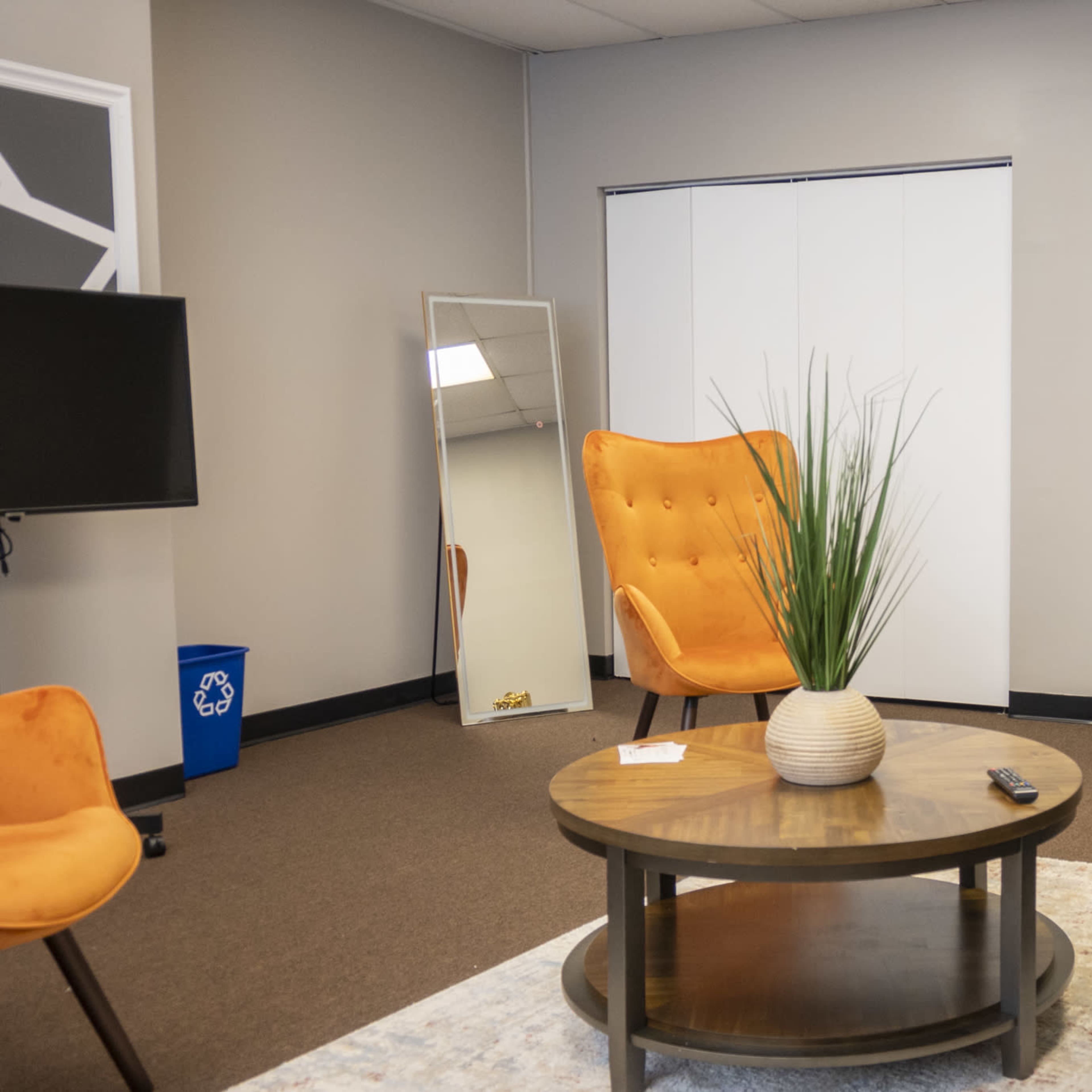 A furnished room with two orange chairs, a gray couch with blue and yellow pillows, a round coffee table, and a large mirror against one wall.