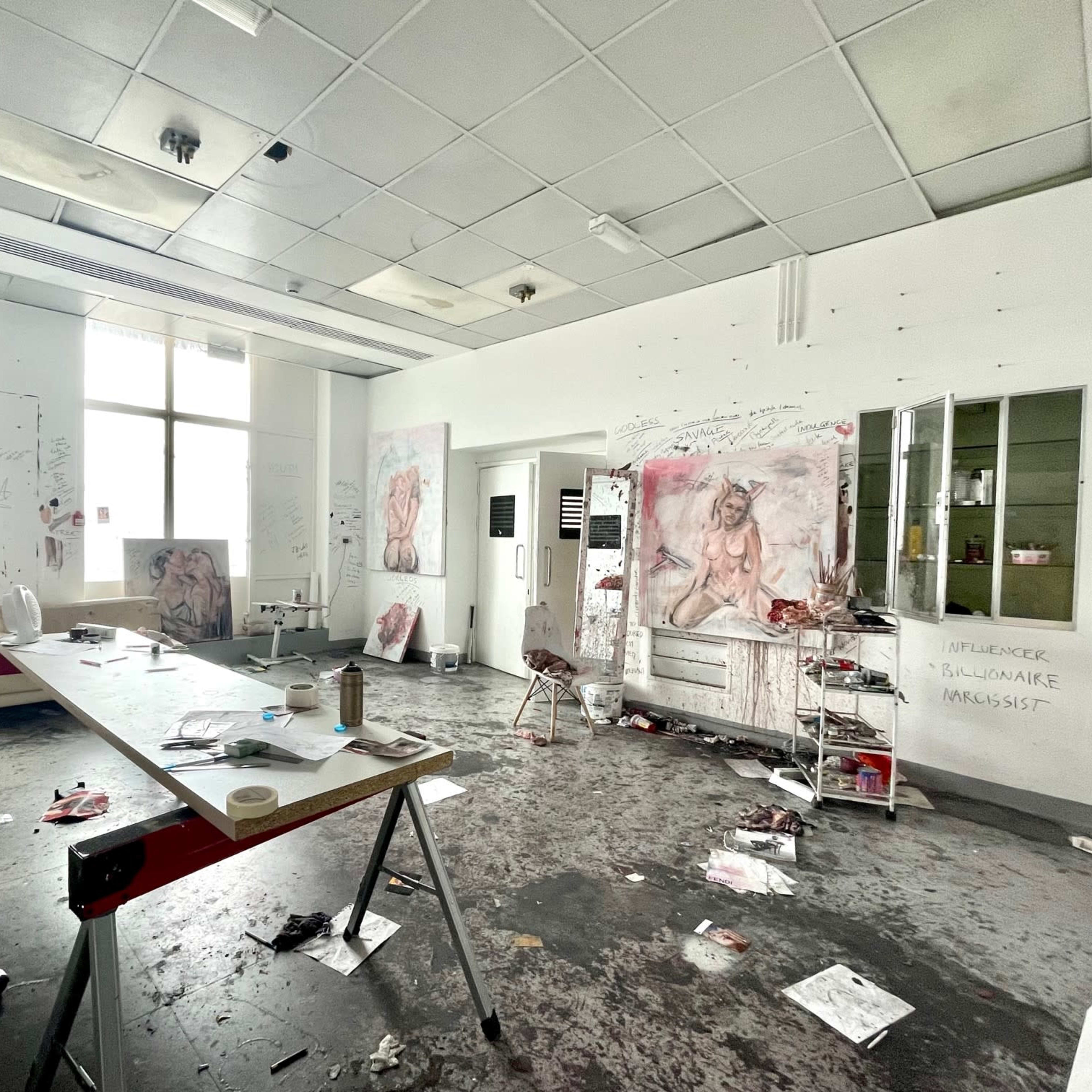 An art studio is cluttered with paint stains and scattered supplies, featuring several paintings on white walls and handwritten notes.