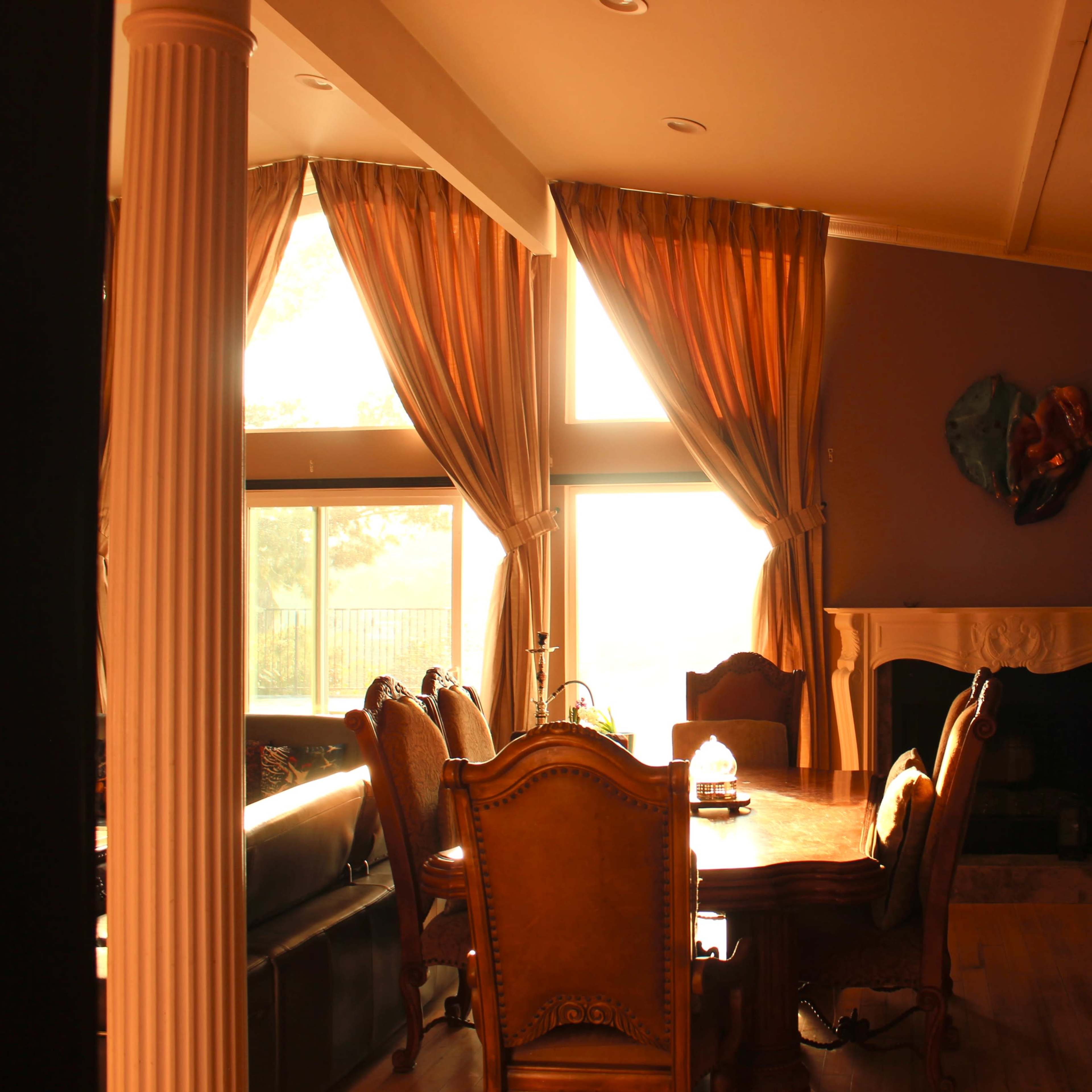 A dining room with a large wooden table surrounded by chairs, illuminated by natural light filtering through tall windows with sheer curtains.