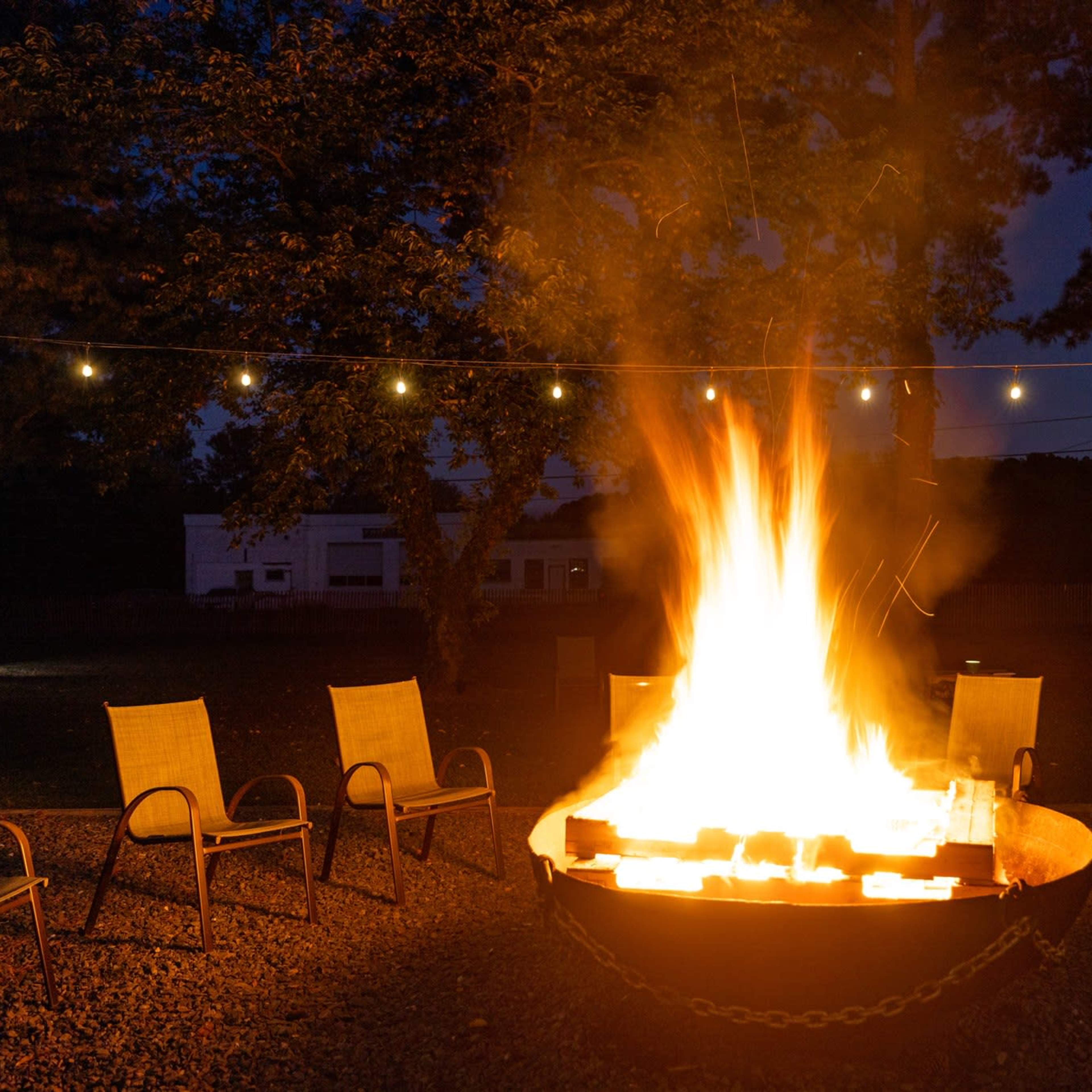 A large fire burns in a metal fire pit surrounded by several empty chairs, with string lights hanging overhead in a darkened outdoor setting.
