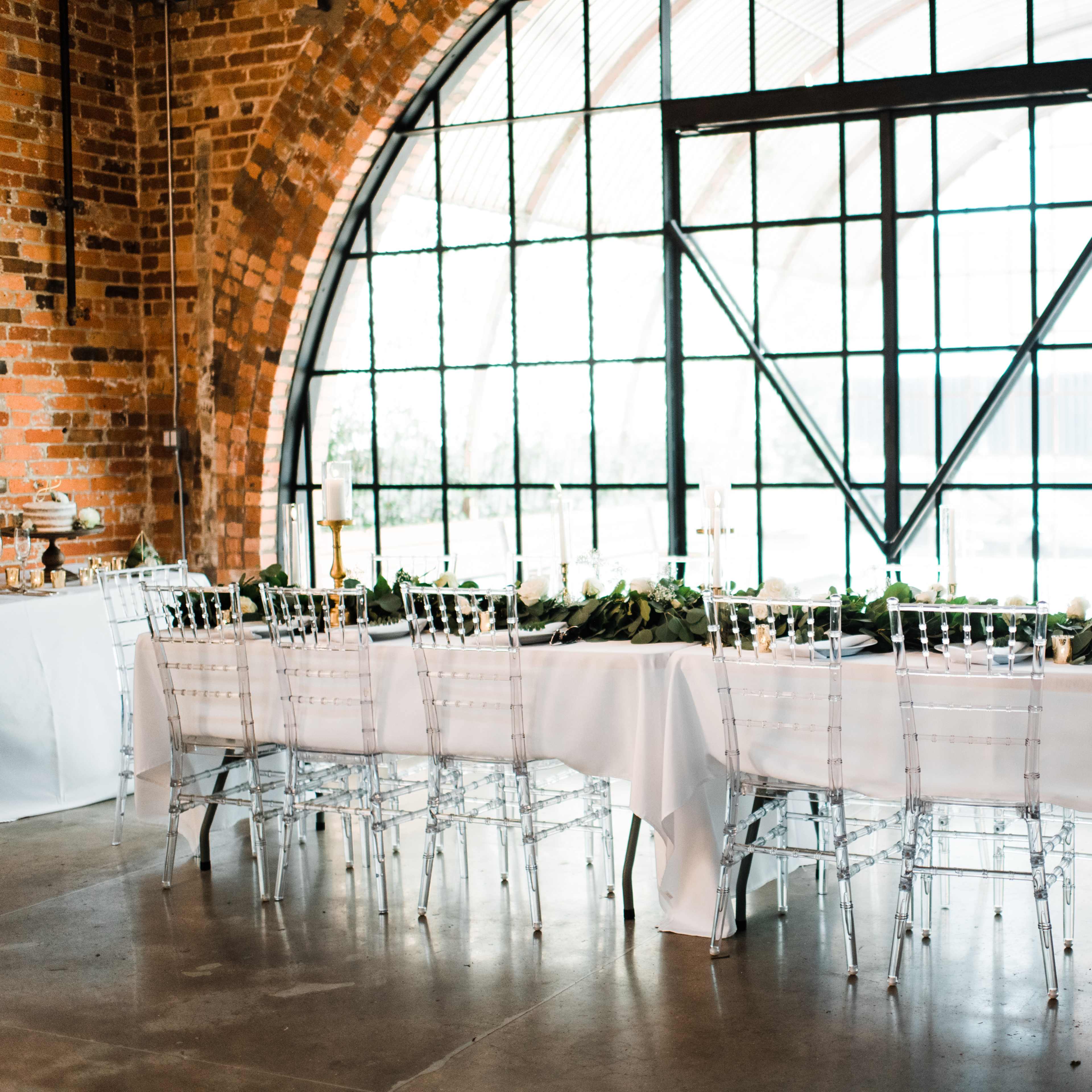 A long dining table adorned with greenery and set with clear chairs is positioned near large arched windows in a brick-walled venue.