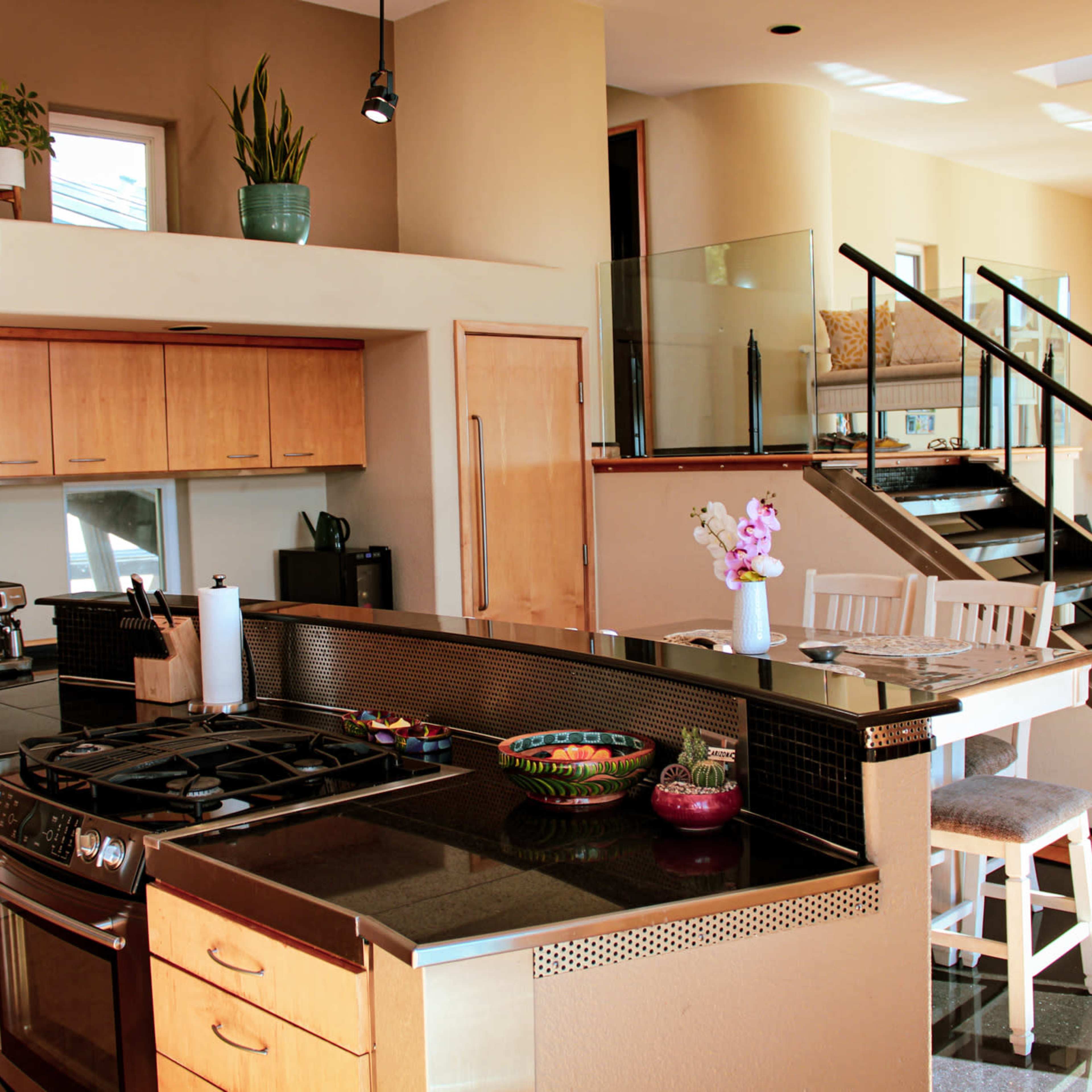 A modern kitchen with wooden cabinets, a large gas stove, and a breakfast bar, featuring an open layout that connects to a living area with a staircase.